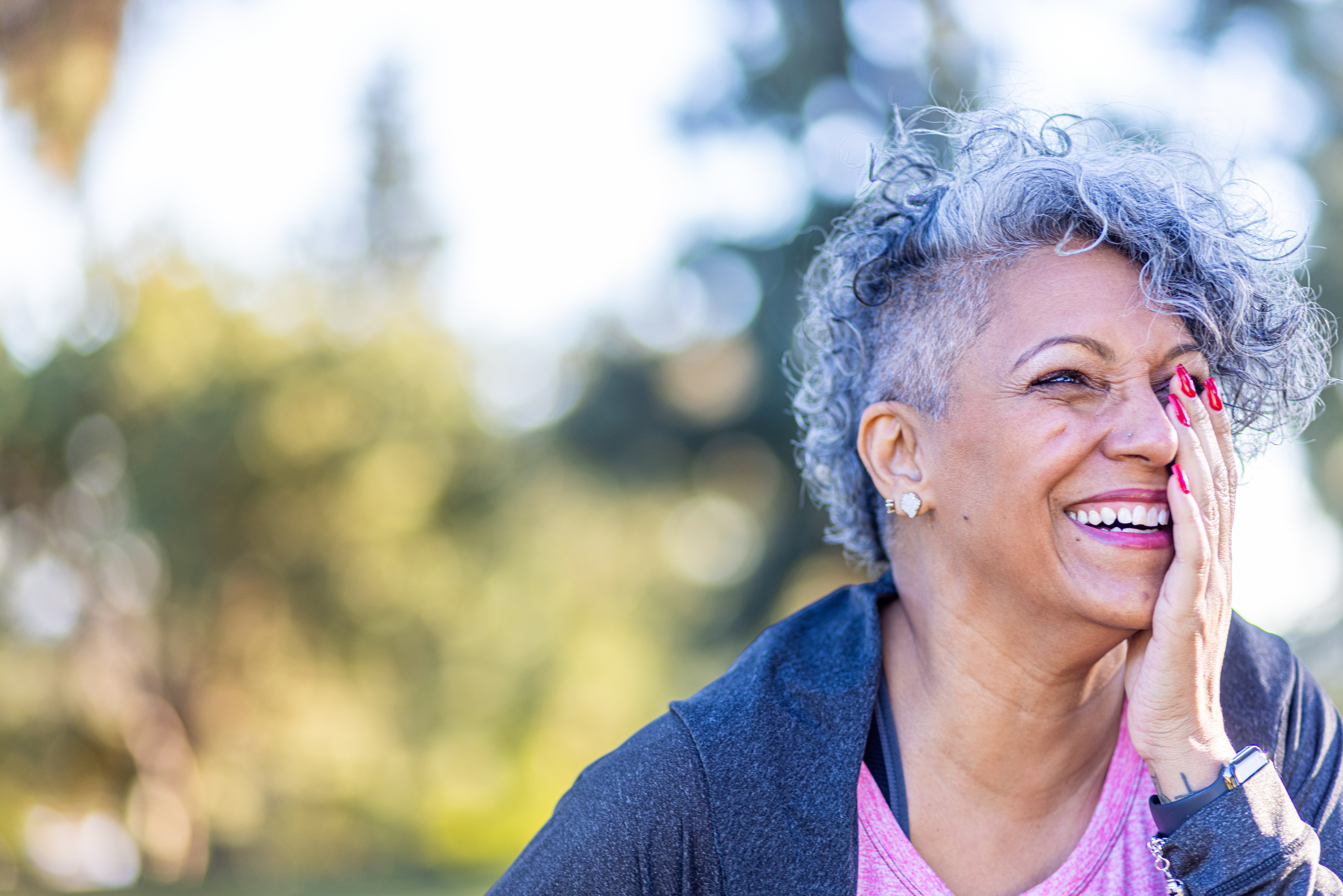 A person with short curly hair is outdoors, smiling, and covering their mouth with one hand, conveying a joyful expression