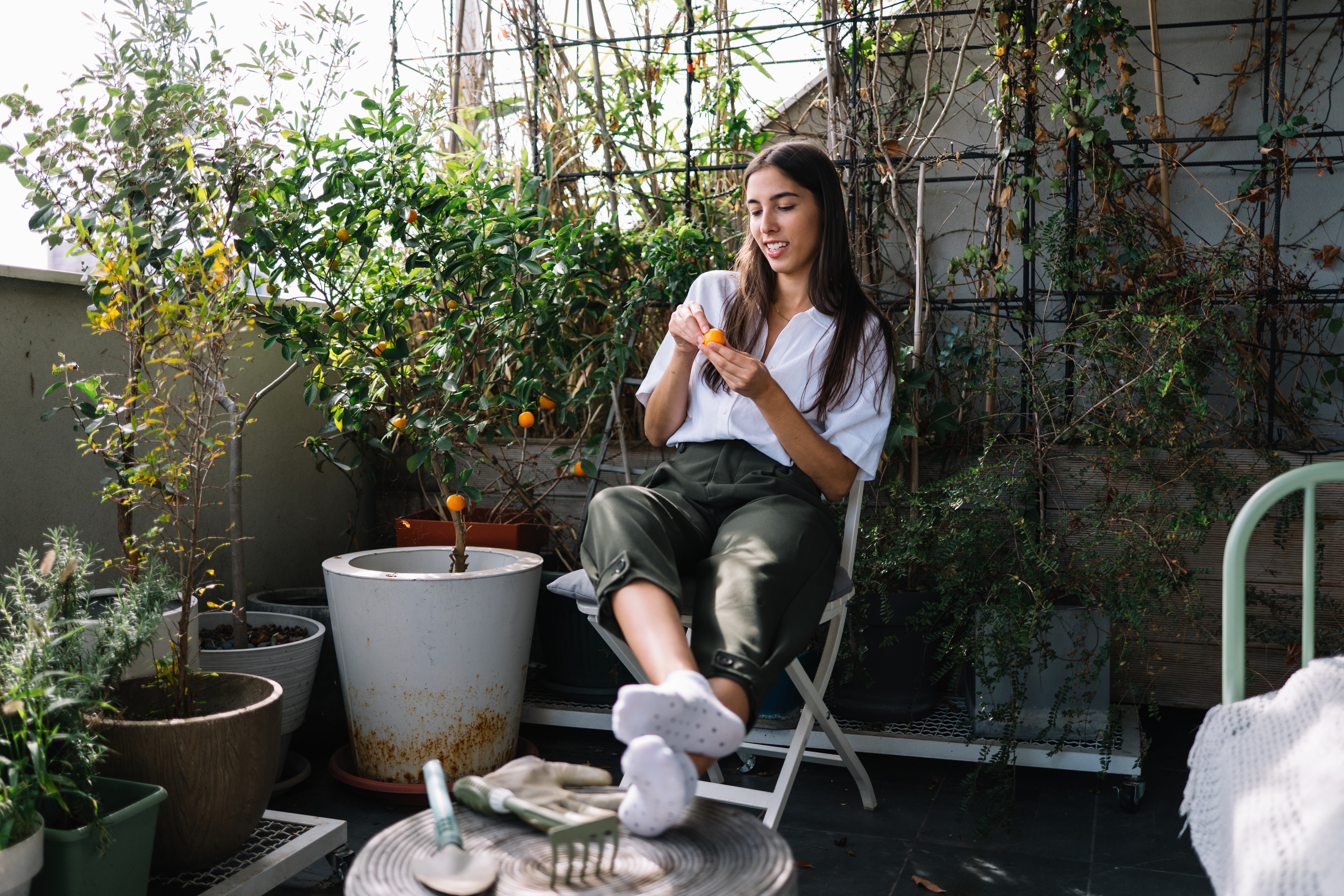 Woman on a balcony tends to plants while seated. She wears a white top and green pants, surrounded by potted greenery