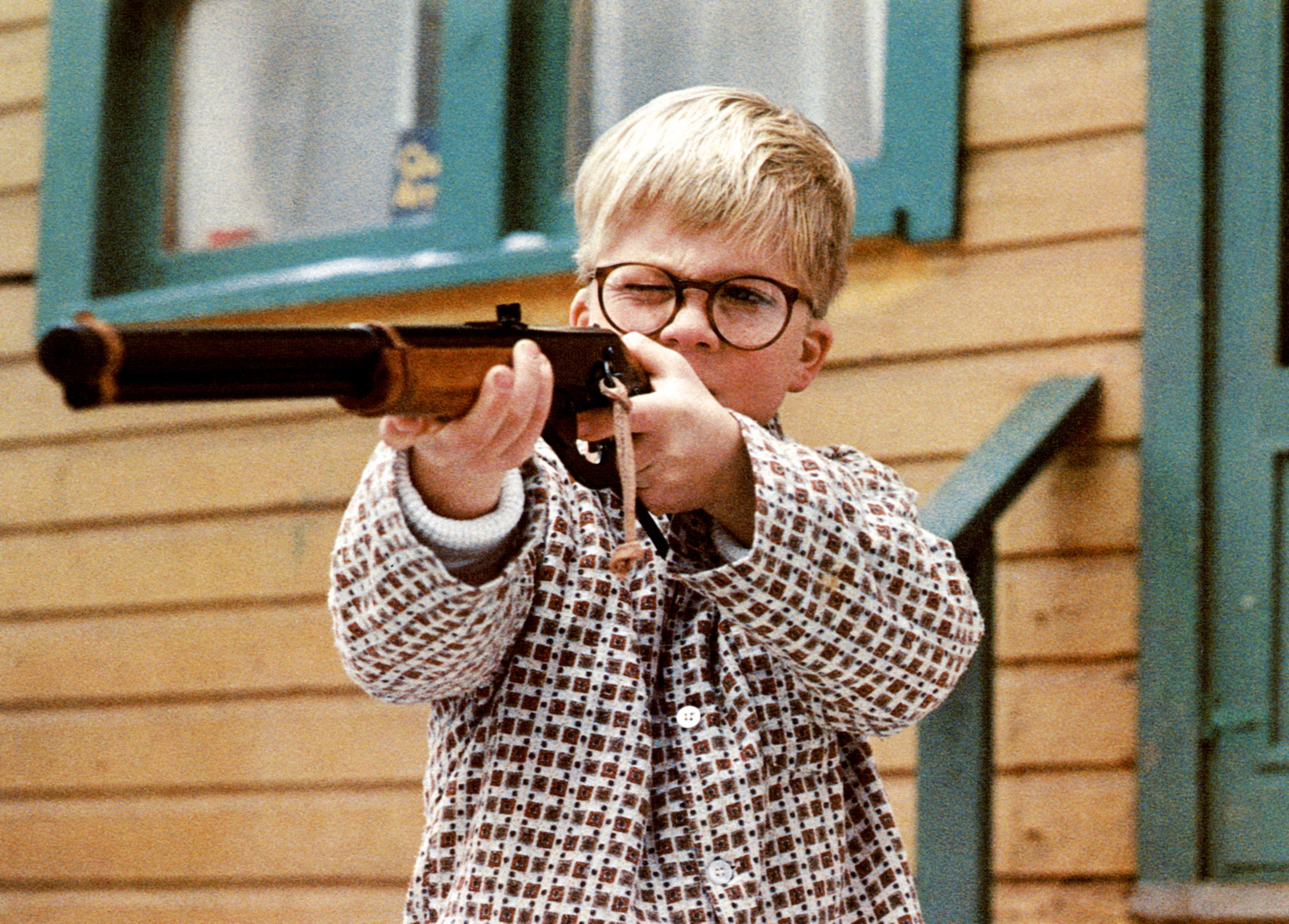 Child aiming a toy gun, wearing glasses and a checkered pattern shirt, standing outside a wooden building