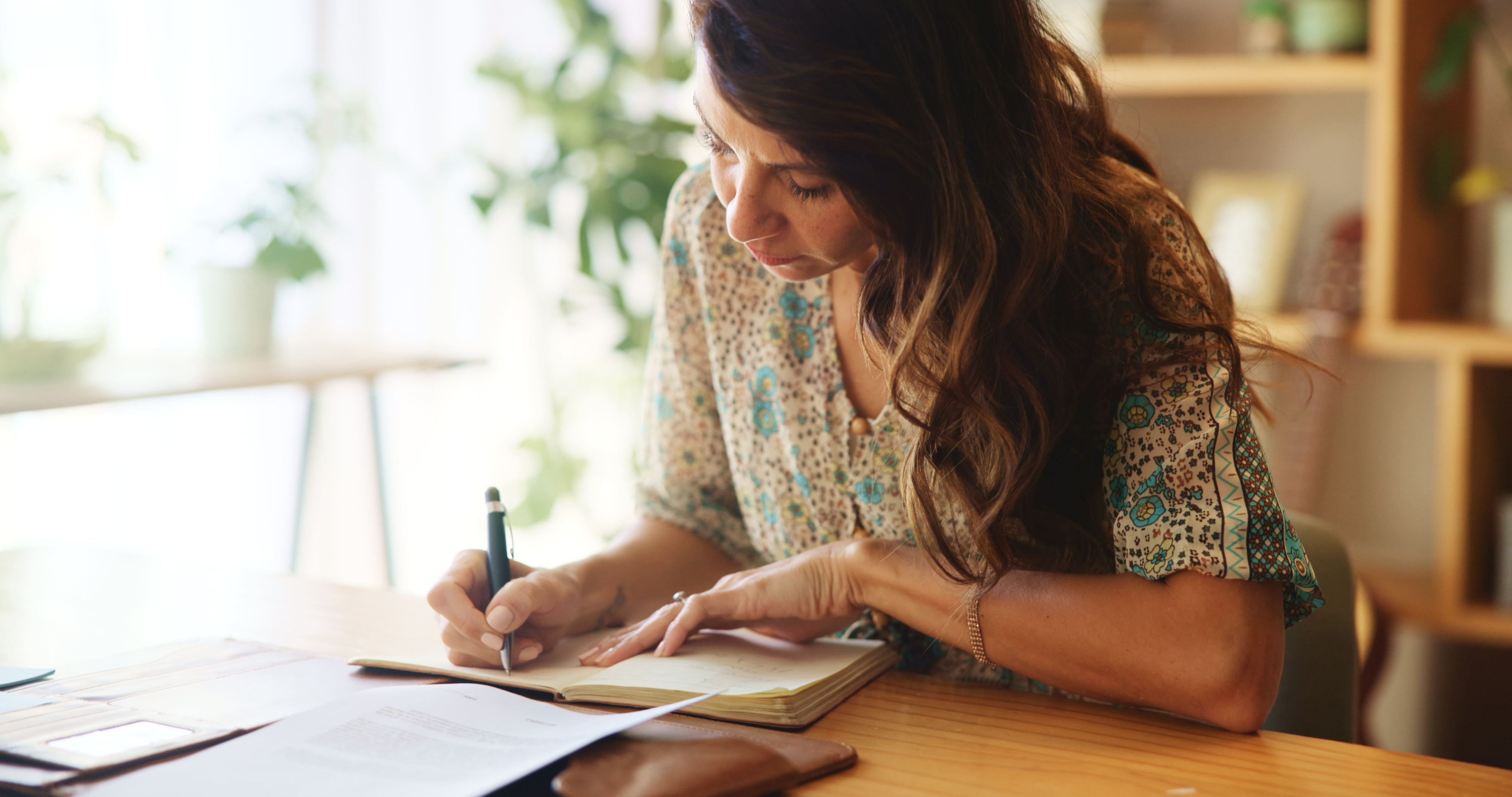 Woman writing in a notebook at a desk, surrounded by documents and plants, conveying a work or financial planning setting