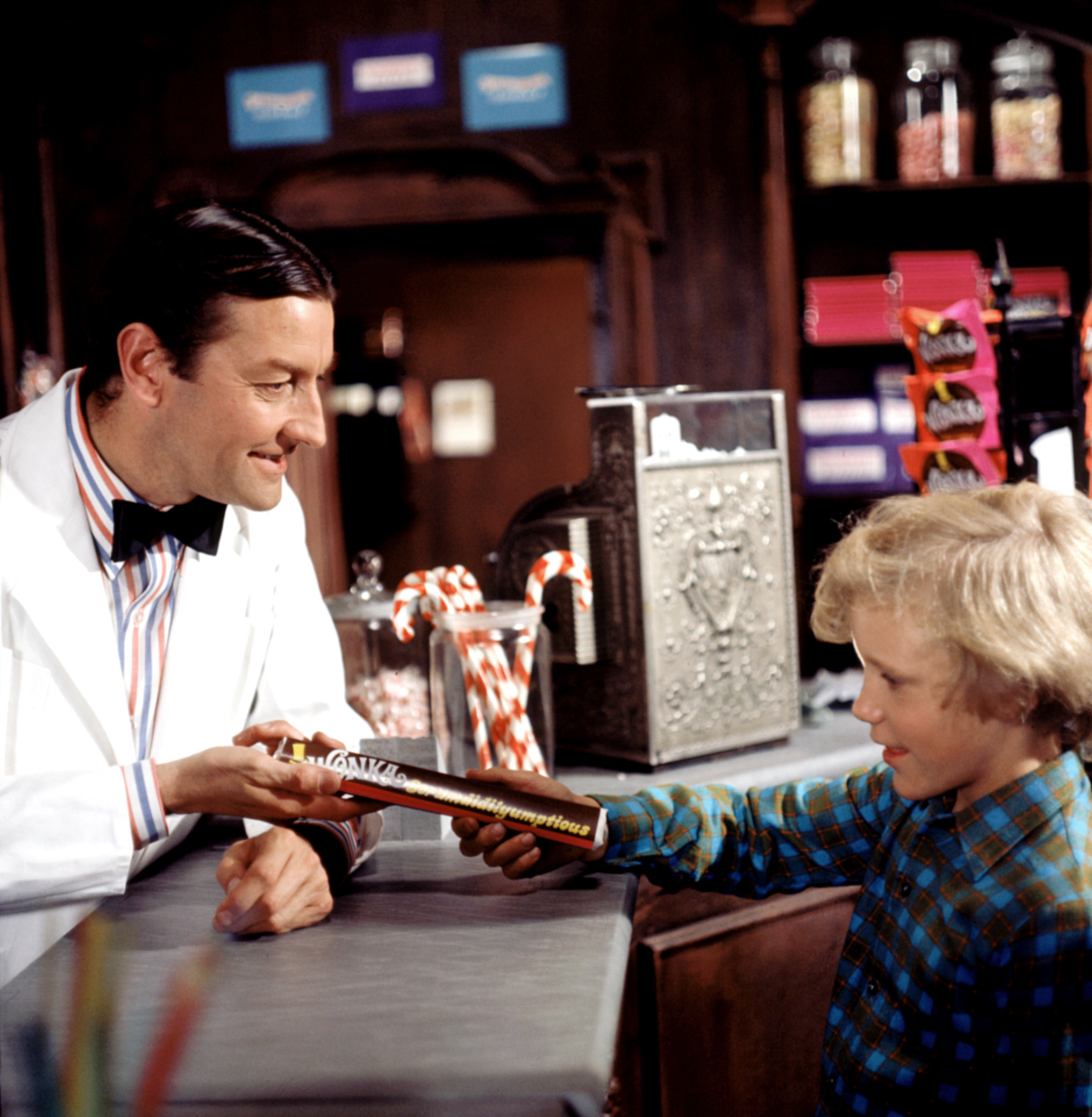 A shopkeeper hands a chocolate bar to a smiling child in a candy store. Shelves in the background display candy jars and boxes