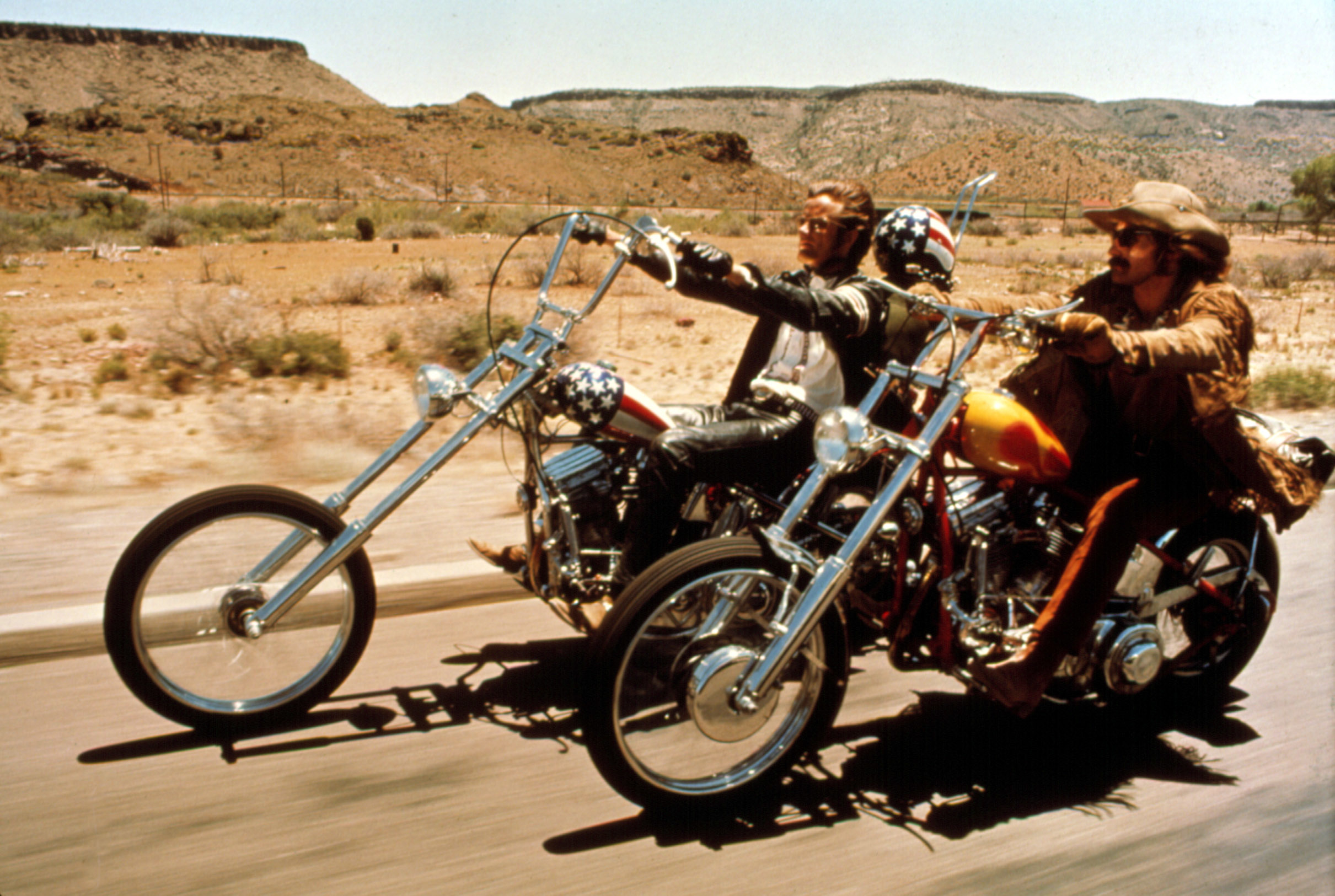 Two people riding chopper motorcycles on a desert highway, wearing casual, rugged attire, with one wearing a helmet