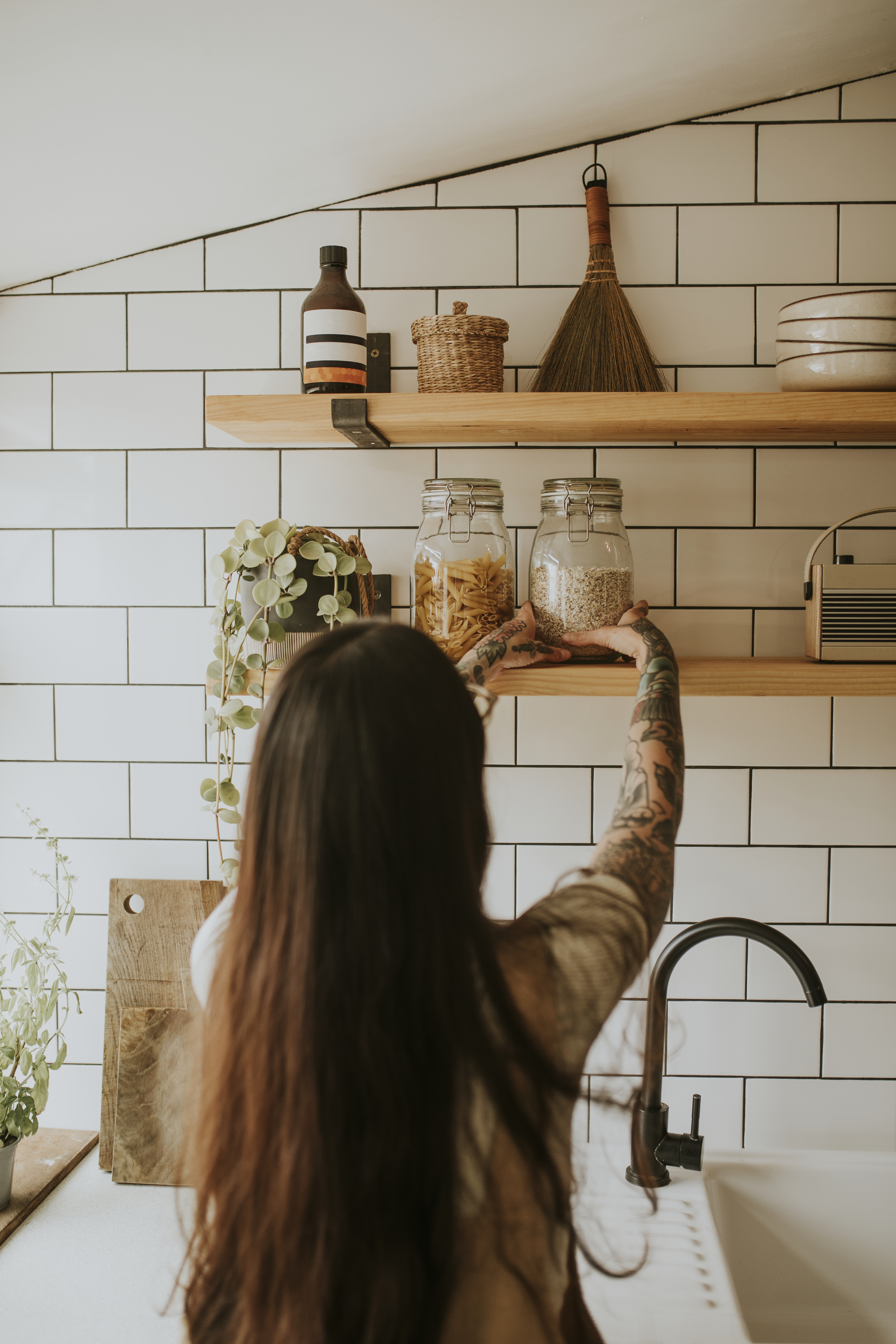 A person with tattoos reaches for jars on a wooden kitchen shelf, surrounded by plants and kitchen items