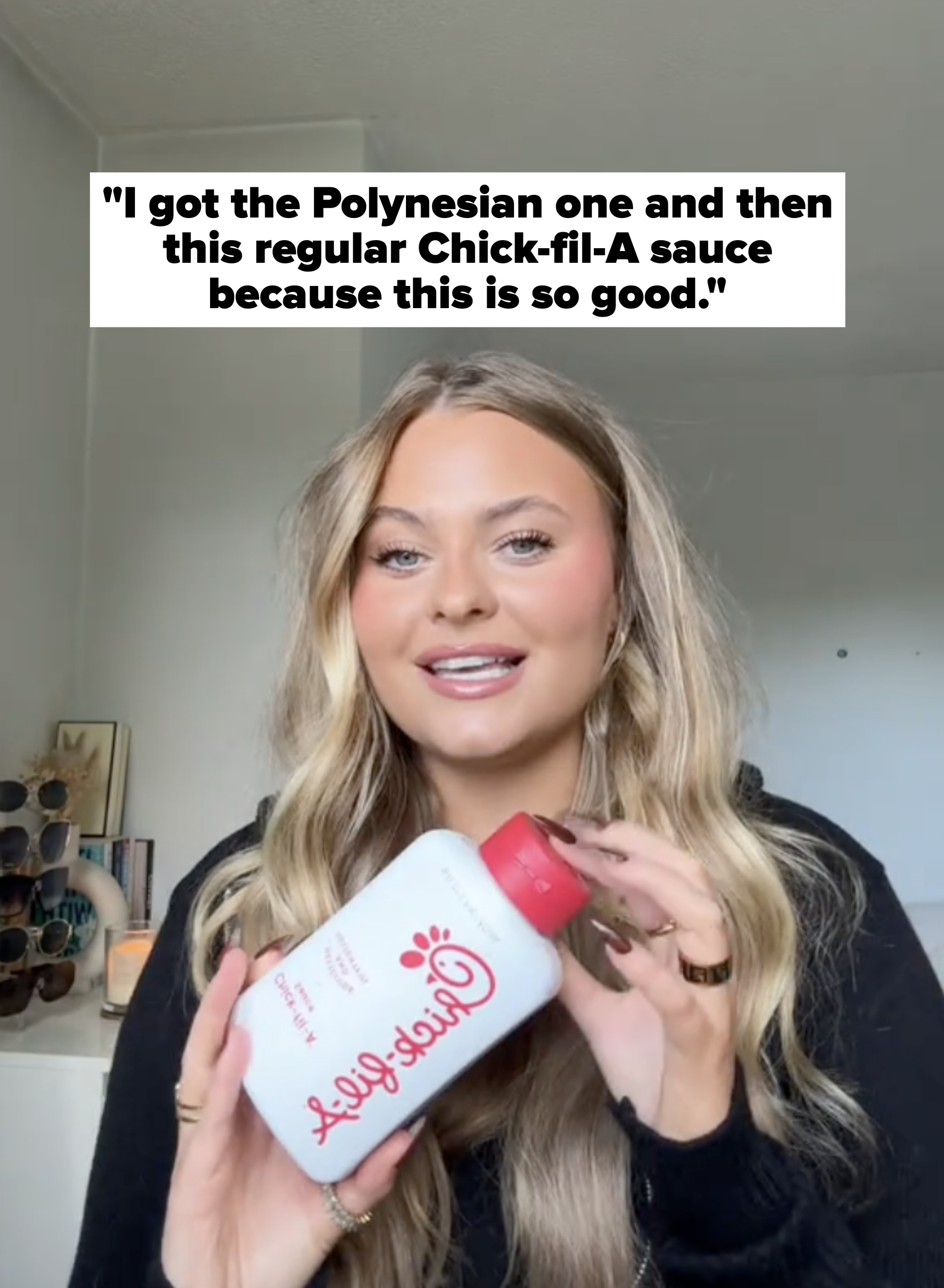 Person smiling and holding a Chick-fil-A sauce bottle. They&#x27;re in a casual setting with sunglasses on a shelf in the background