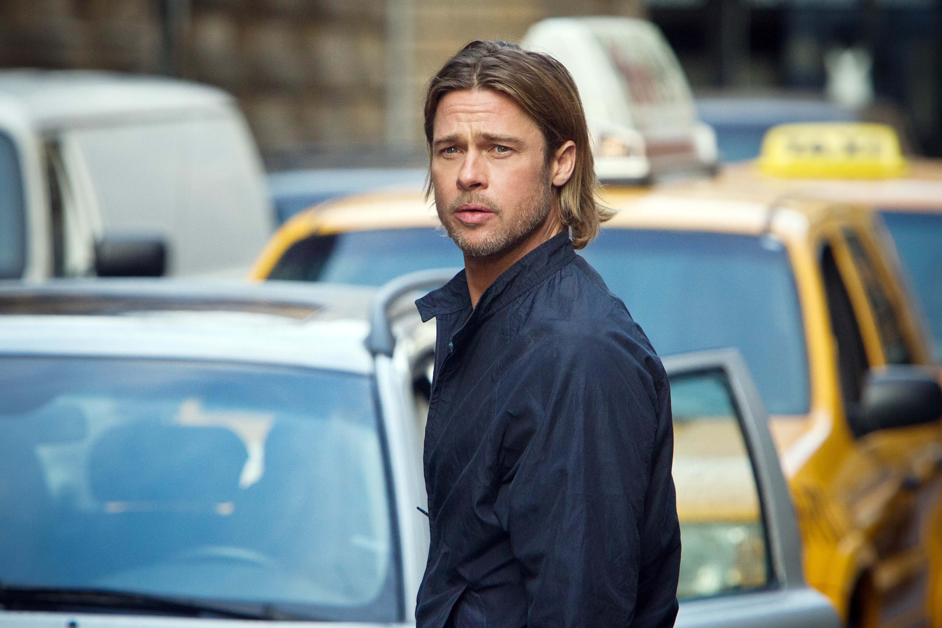 Person with shoulder-length hair in a city street, wearing a casual jacket, taxis in background