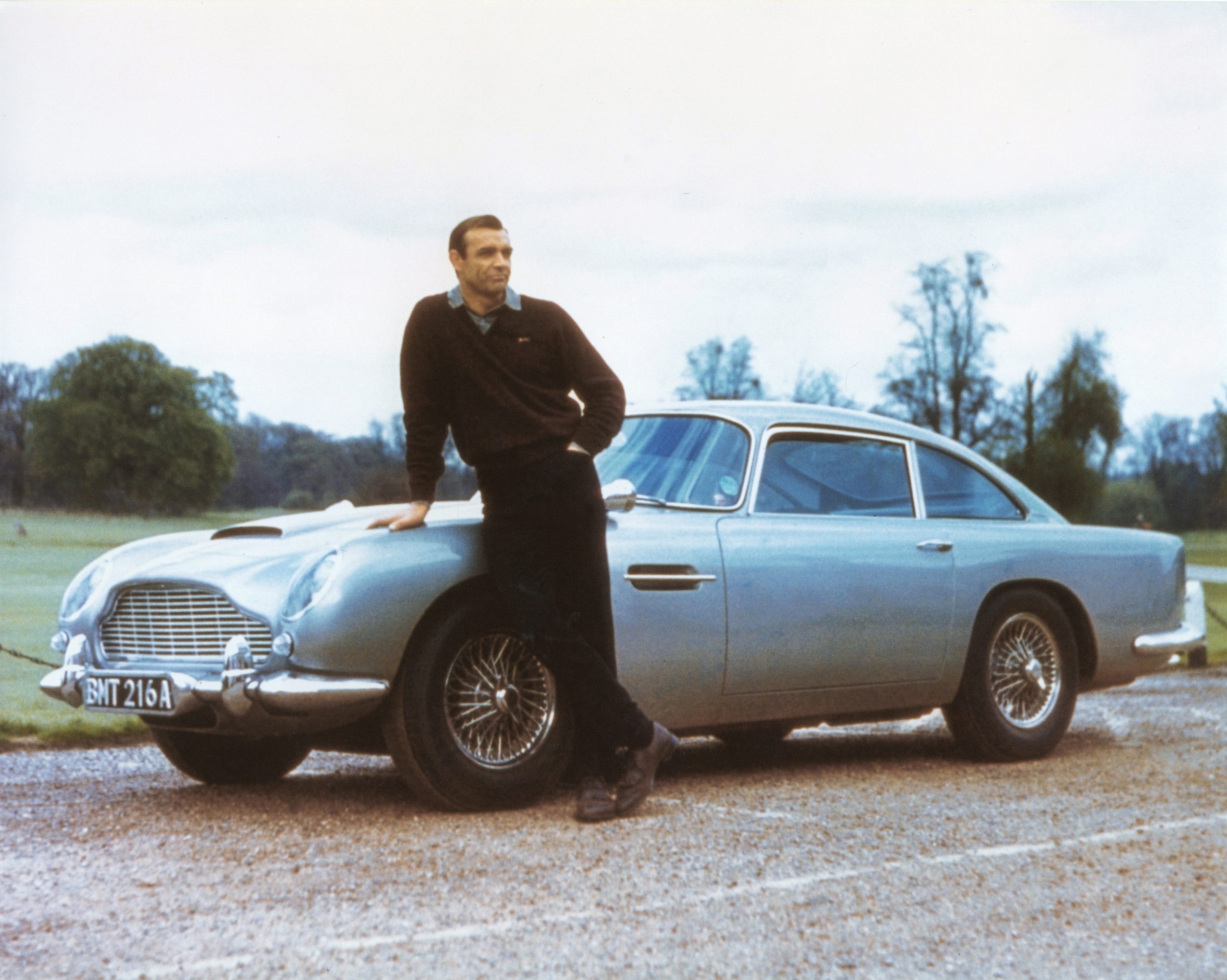 Person leaning against a classic car on a country road, wearing casual attire, in a scene associated with TV and movies