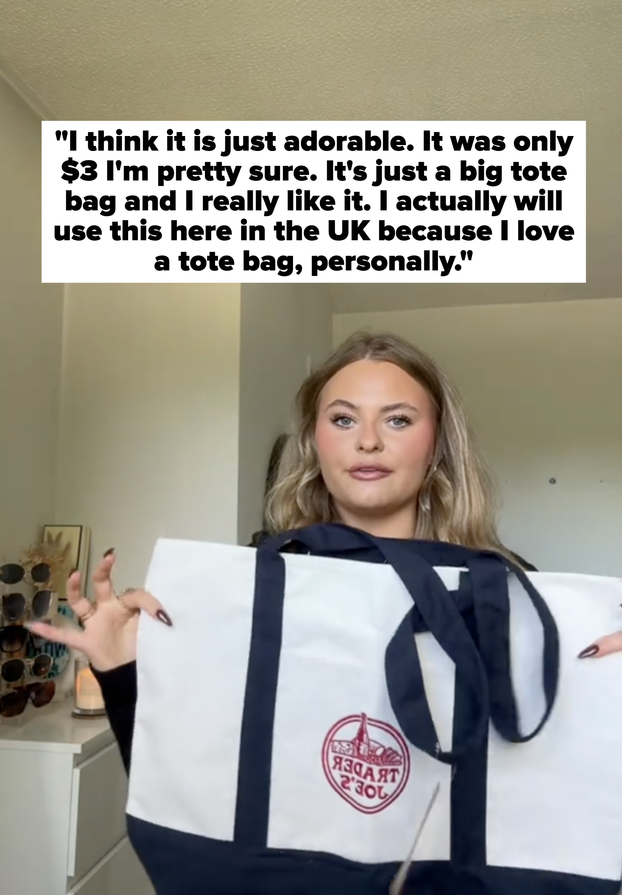 A person holds up a Trader Joe&#x27;s tote bag while standing in a room with sunglasses displayed on a dresser