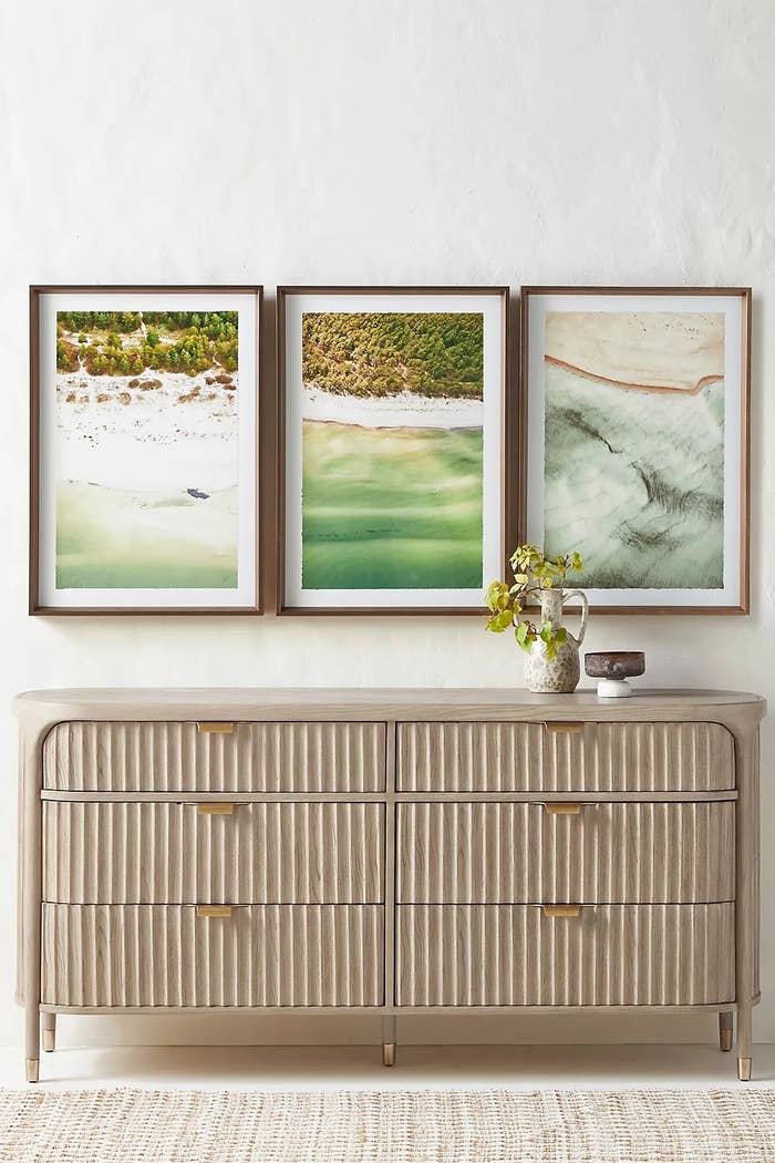 Modern dresser with gold accents, topped with decorative items, beneath three nature-themed framed artworks