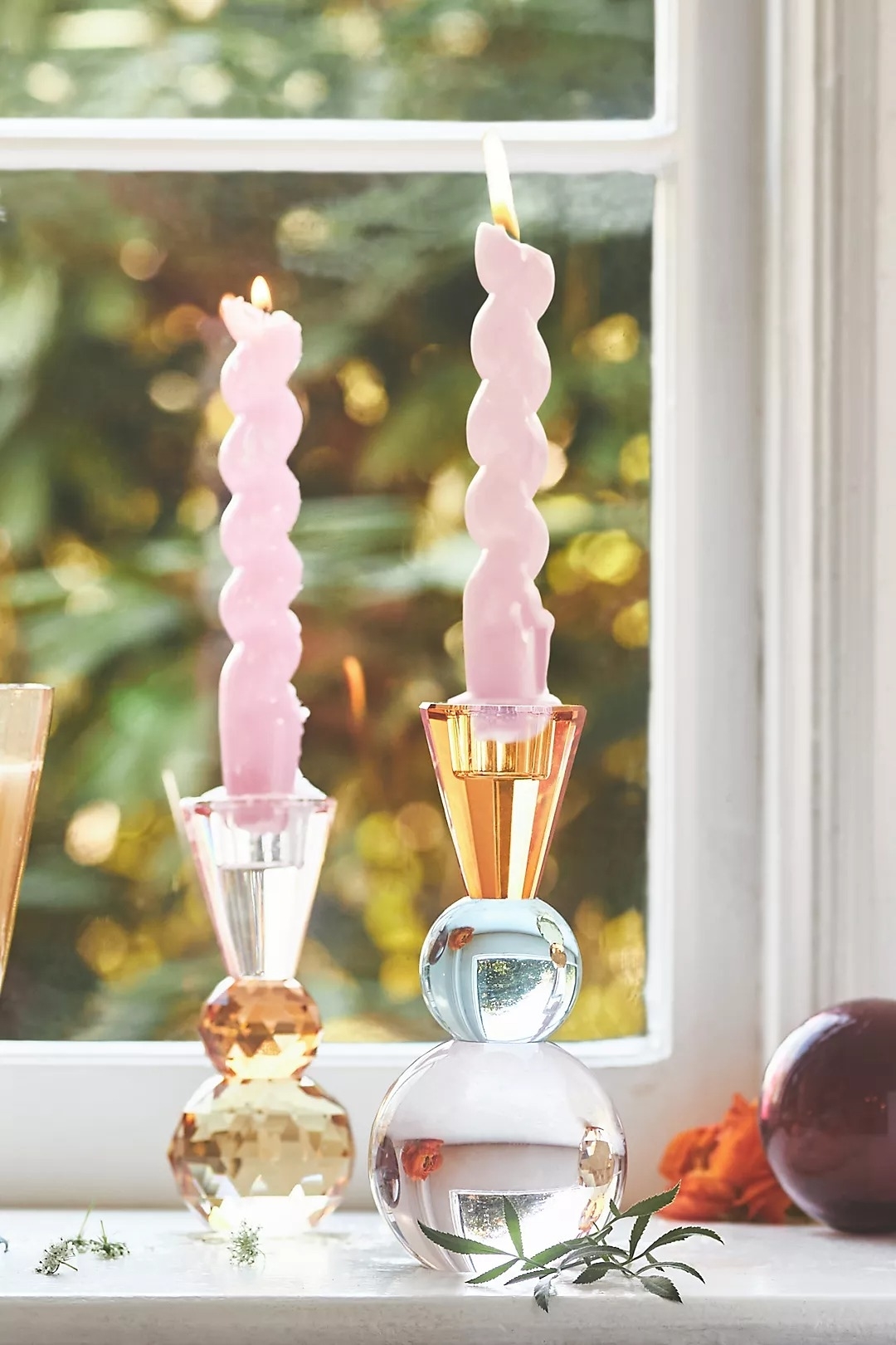 Two pink spiral candles on geometric glass holders by a window, with greenery outside. Suitable for home decor shopping