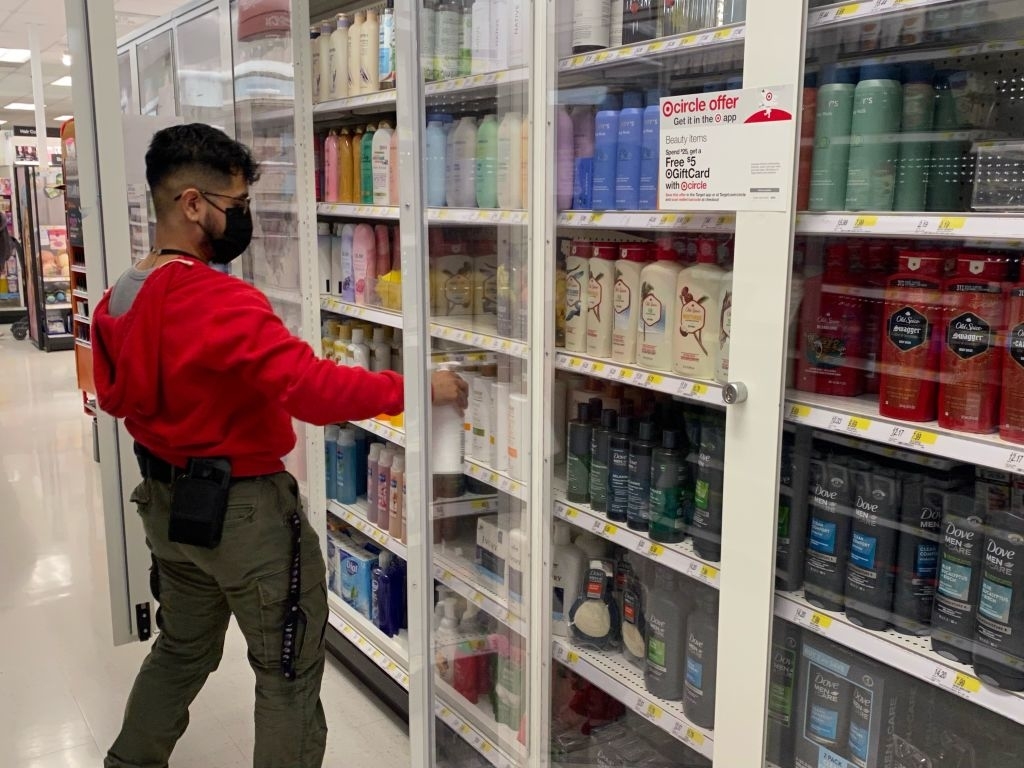 Person shopping in a store aisle with personal care products, wearing a mask, red hoodie, and pants. Shelves are behind locked glass doors