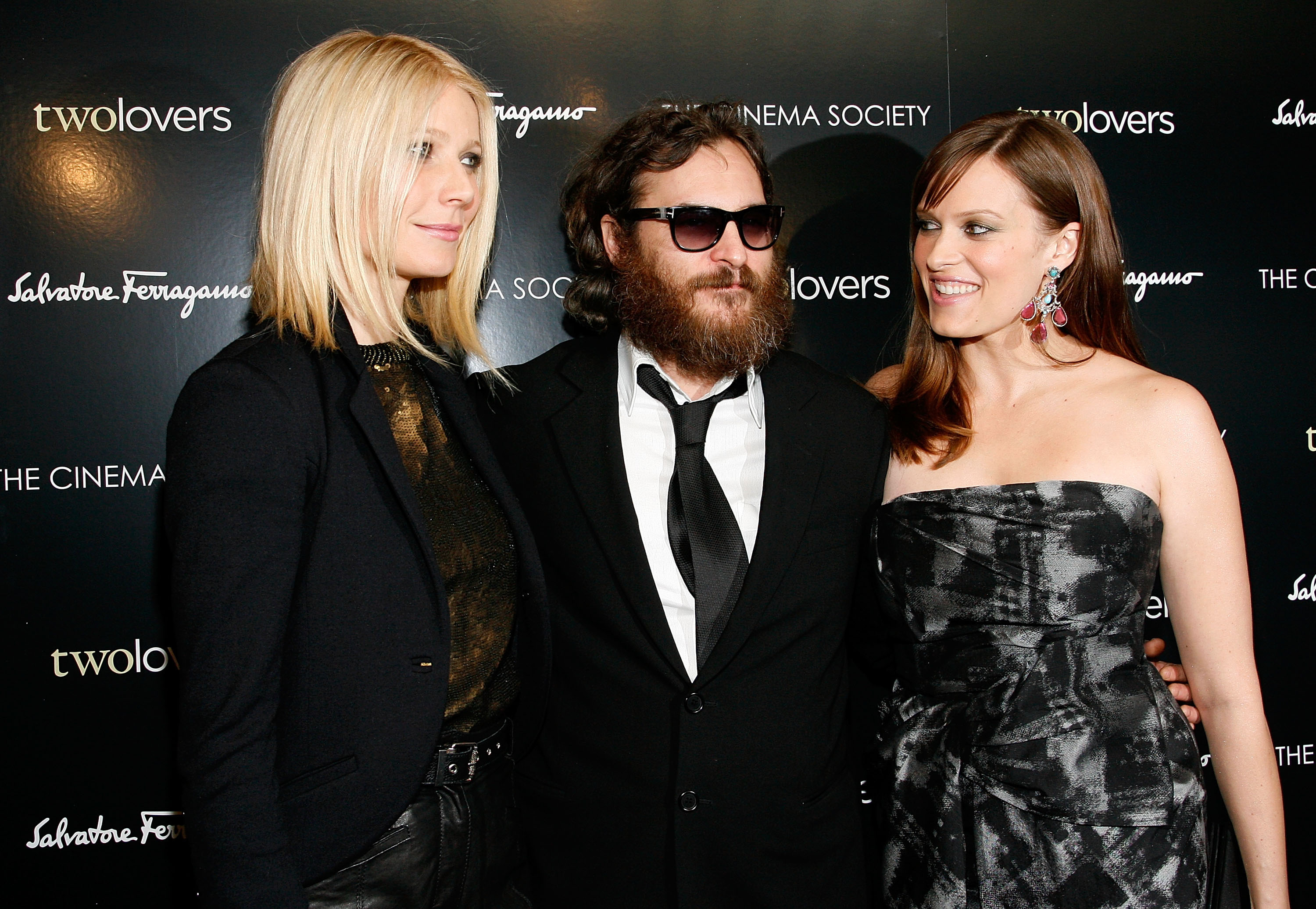 Three people pose at a &quot;Two Lovers&quot; event. The person on the left wears a blazer, the center person a suit and sunglasses, and the right person a strapless dress