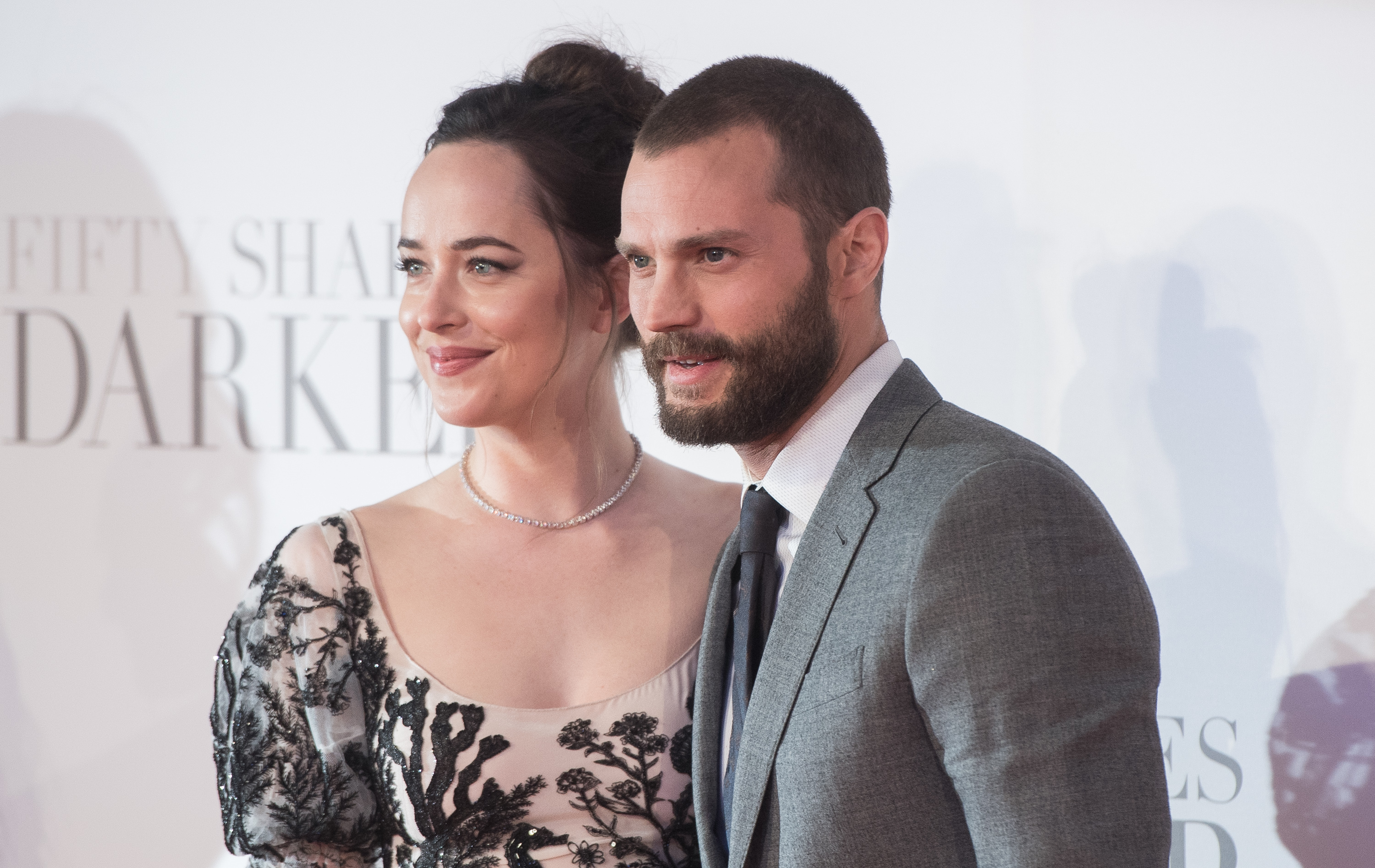 Two people pose on the red carpet; one wears a floral gown, the other a gray suit with a tie