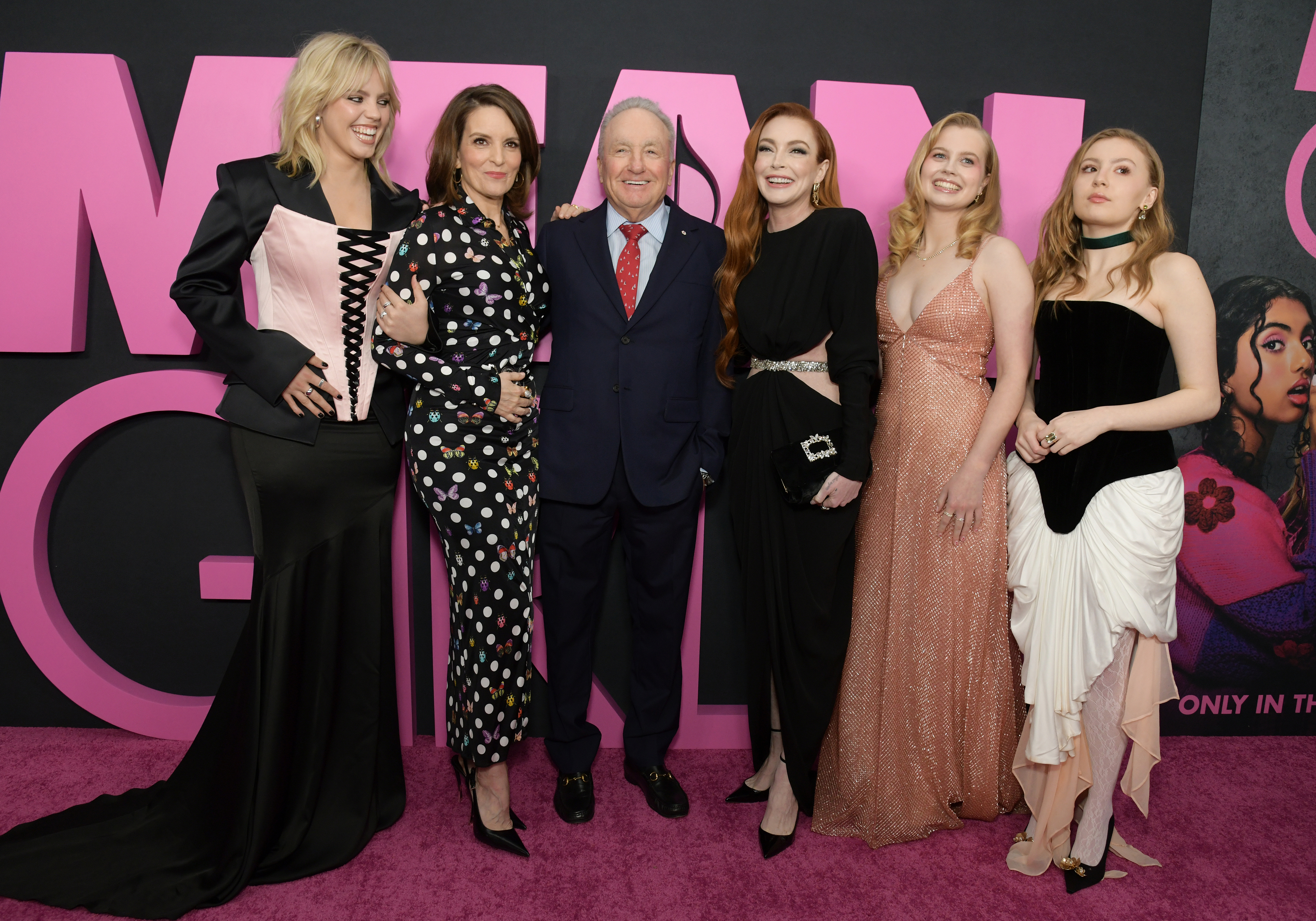 Group of six people posing on a red carpet event with a &quot;Mean Girls&quot; backdrop. Mixed styles: formal suits, elegant dresses, polka dot and corset details