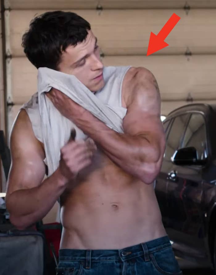 A person in a garage setting wipes their face with a cloth, displaying muscular arms and torso