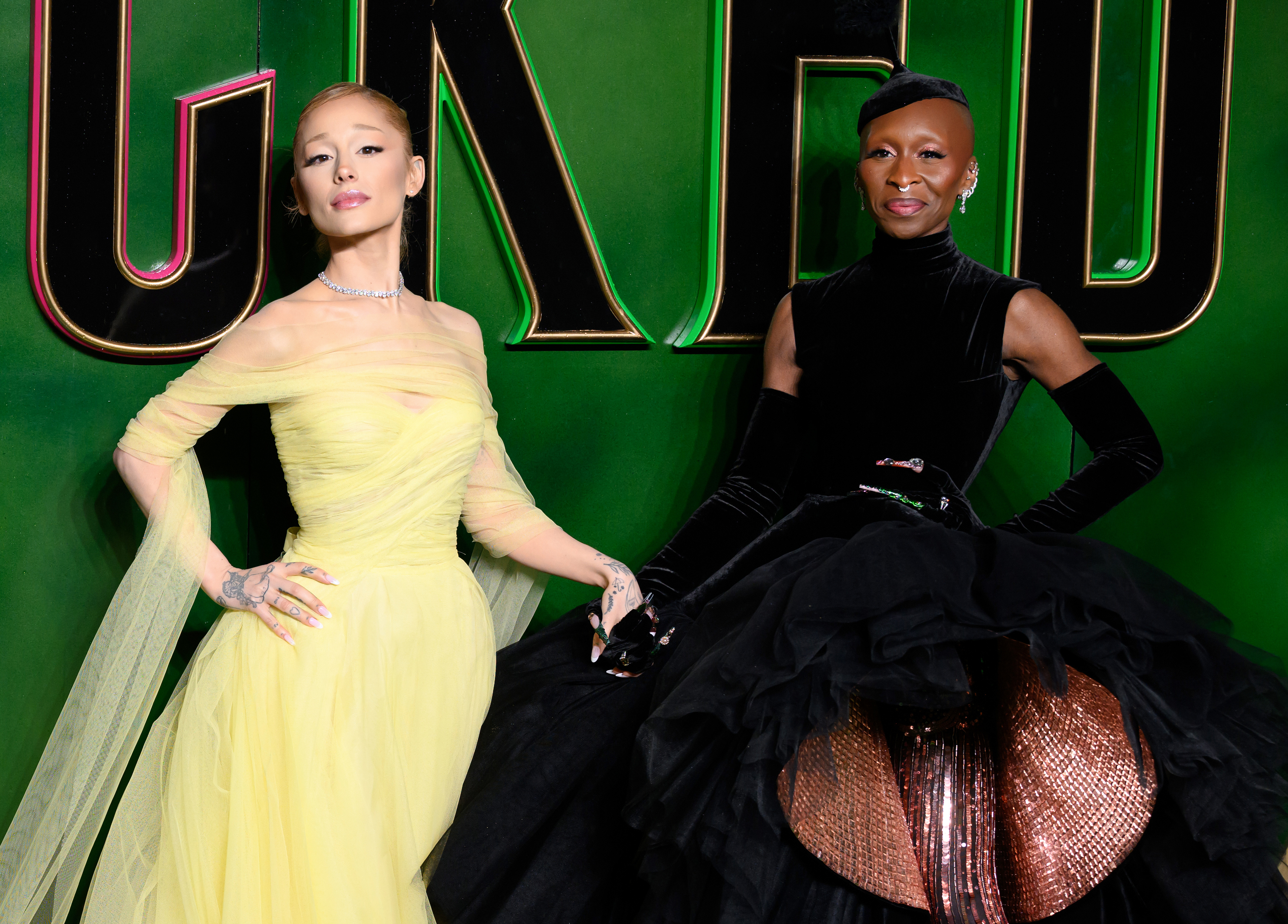 Two individuals pose on a red carpet. The first wears an off-shoulder gown, and the second wears a high-neck, sleeveless dress with gloves and an elaborate skirt