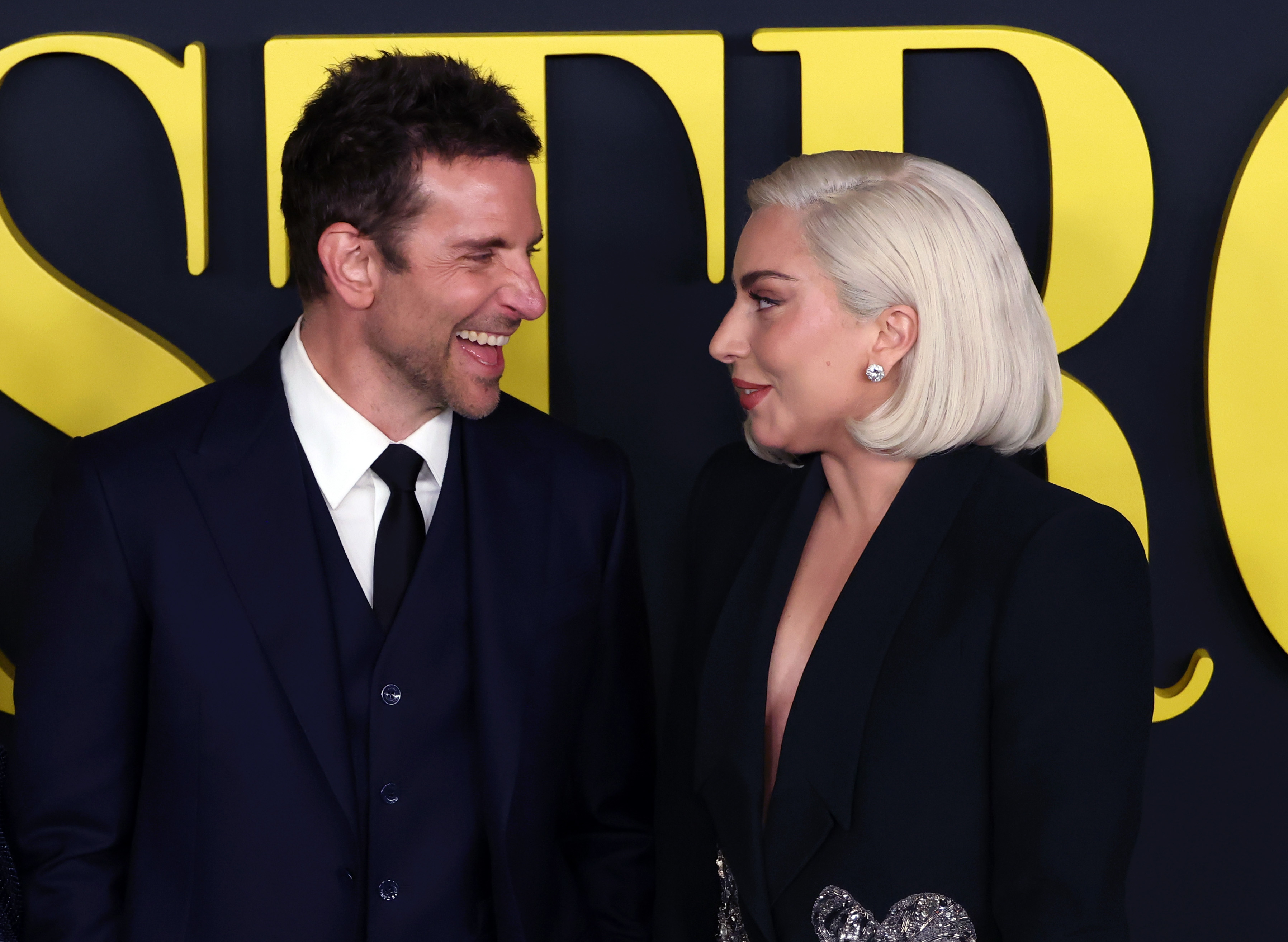 Two people smiling at each other on a red carpet, both wearing elegant, dark outfits against a backdrop with large yellow text