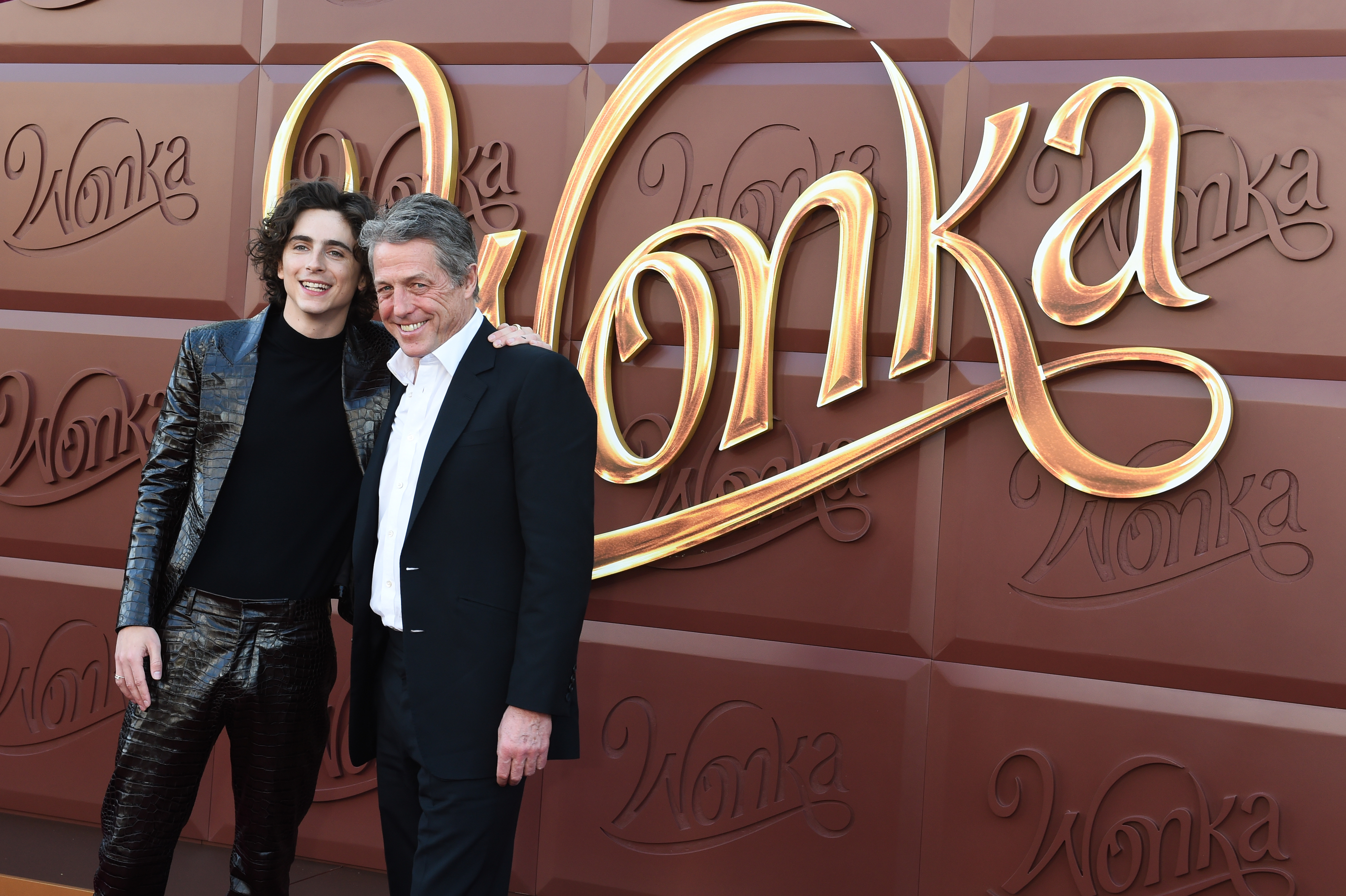 Two people pose at the &quot;Wonka&quot; premiere. One wears a shiny suit, the other in a traditional suit. They stand in front of a background with &quot;Wonka&quot; text