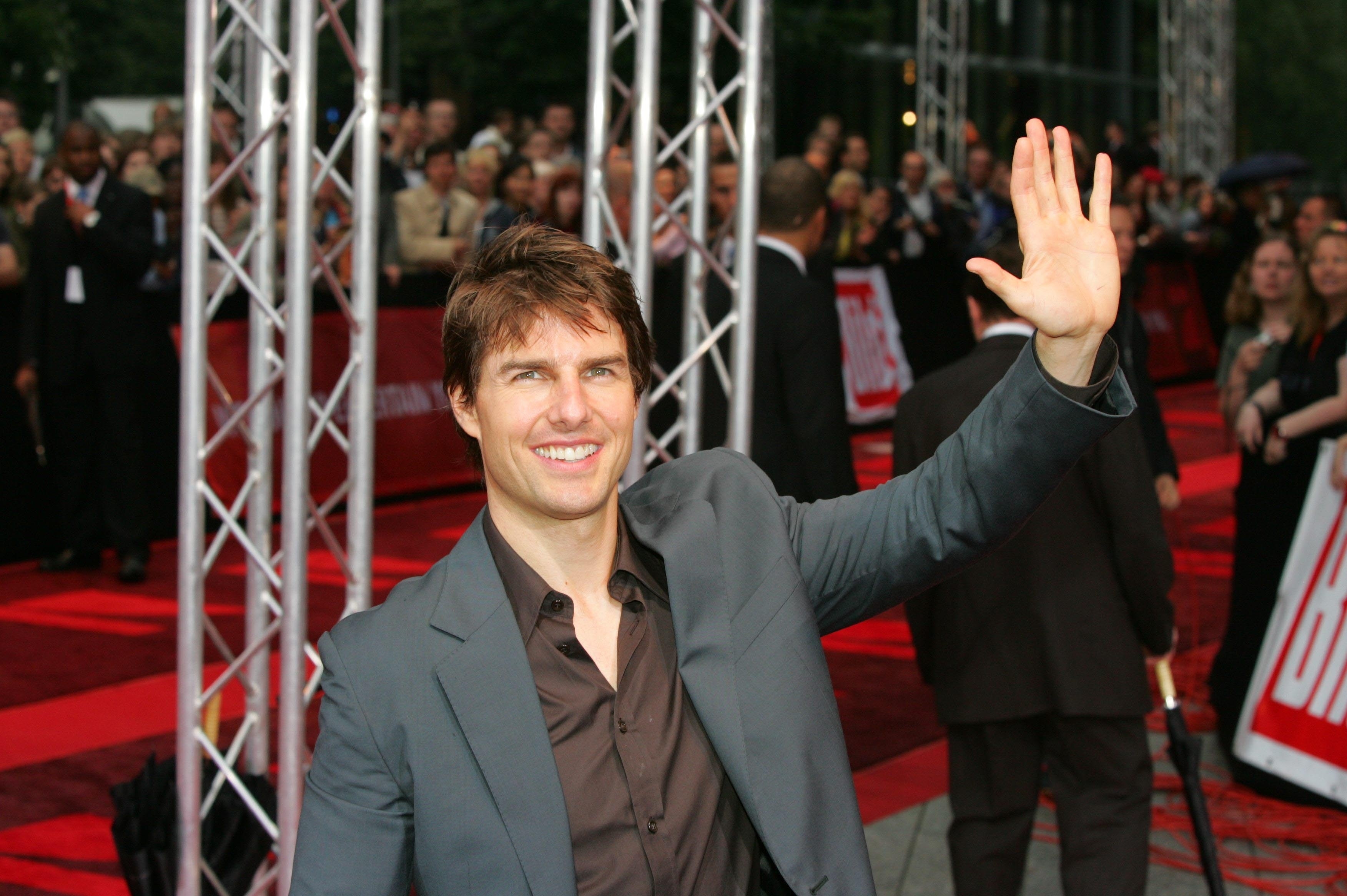 Person in a suit waves on a red carpet, surrounded by a crowd and event staff