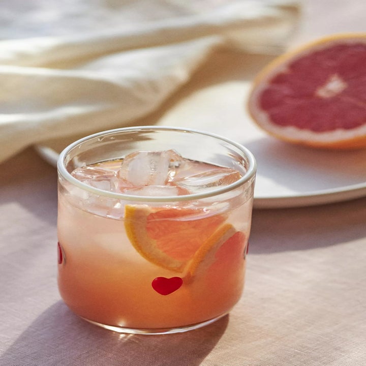A refreshing glass of ice-filled grapefruit juice with heart designs, next to a grapefruit slice on a plate, set on a light tablecloth
