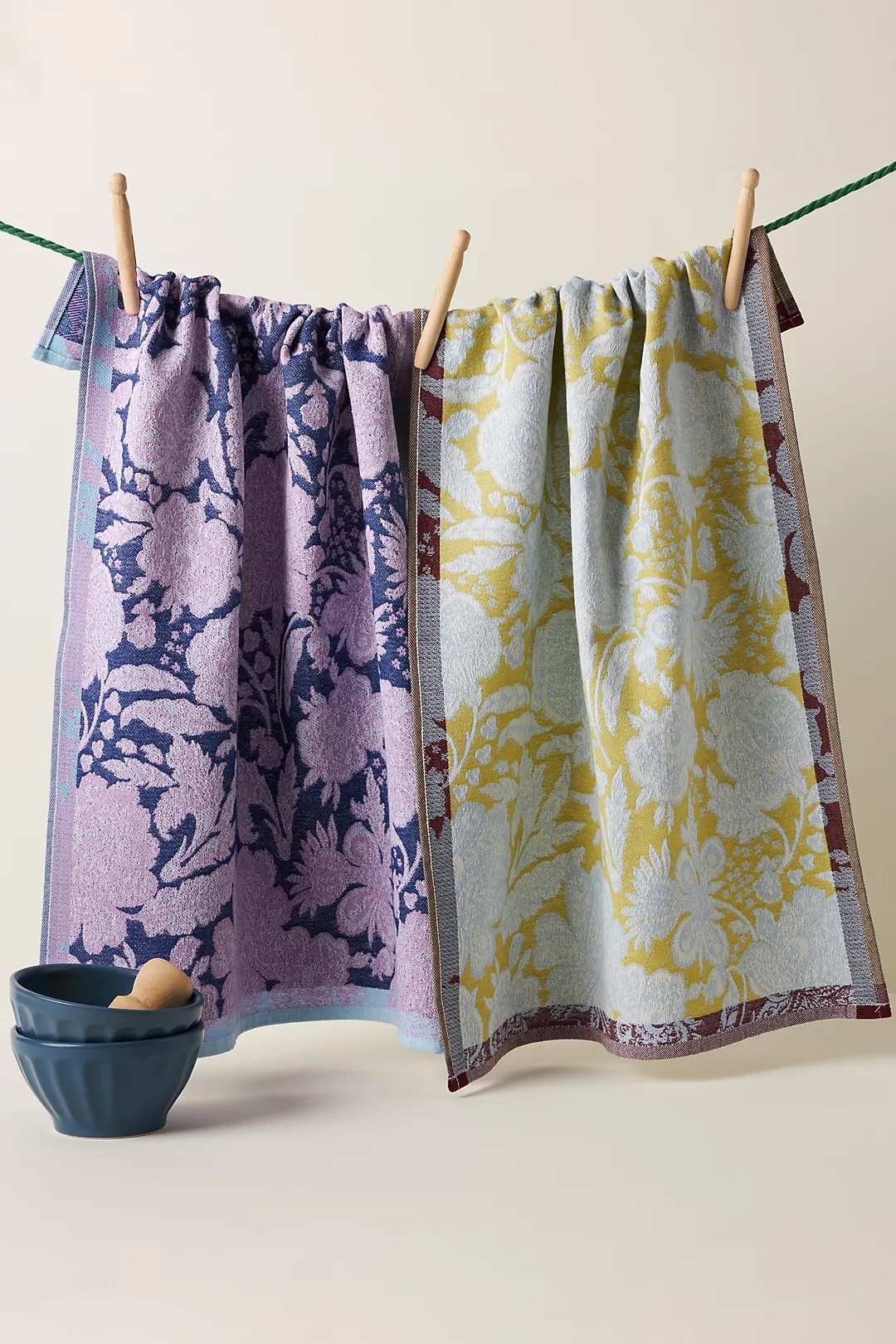 Two decorative towels with floral patterns hang on a line beside a small blue bowl, suitable for kitchen or bathroom decor