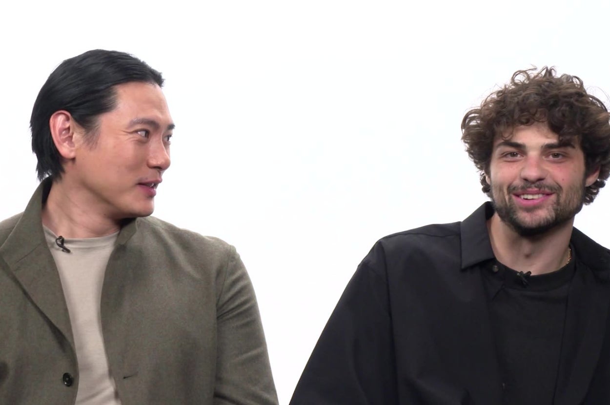 Two men in semi-formal attire are seated, conversing in a studio setting. One wears a blazer, the other a casual jacket