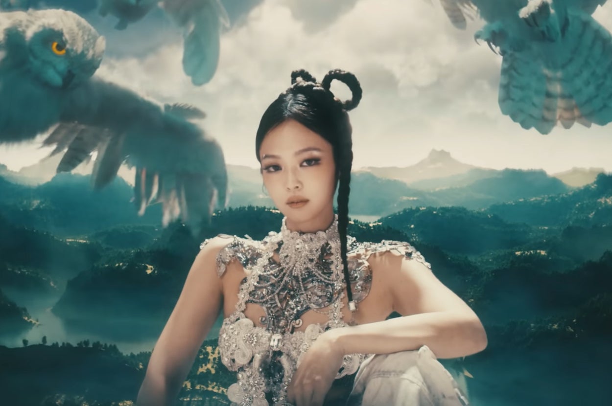 Jennie in elaborate jeweled attire poses with flying owls in a fantasy landscape