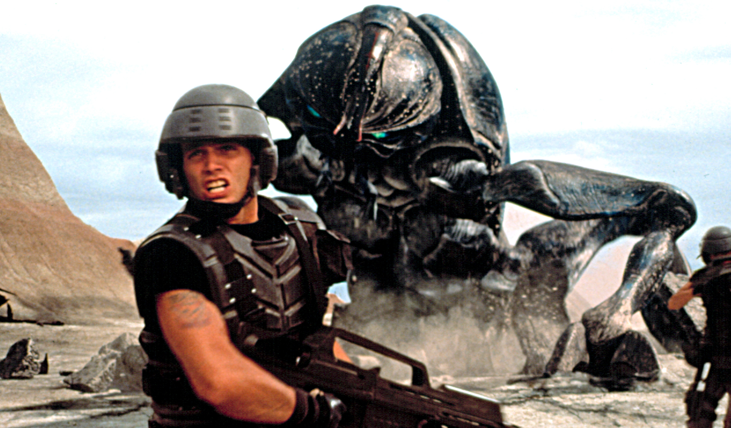 A soldier in futuristic armor faces a large alien insect creature on a rocky terrain, holding a weapon defensively