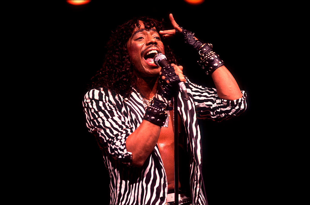 Singer performing energetically connected stage, wearing a bold, striped outfit with studded leather gloves and bracelets, singing into a microphone