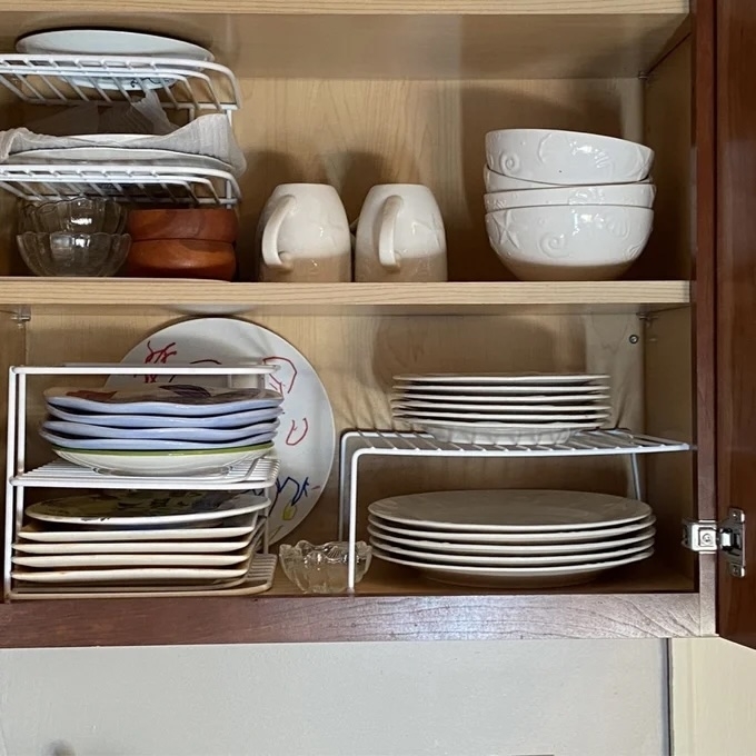 Neatly organized kitchen cabinet with stacked plates, bowls, and mugs on wire racks