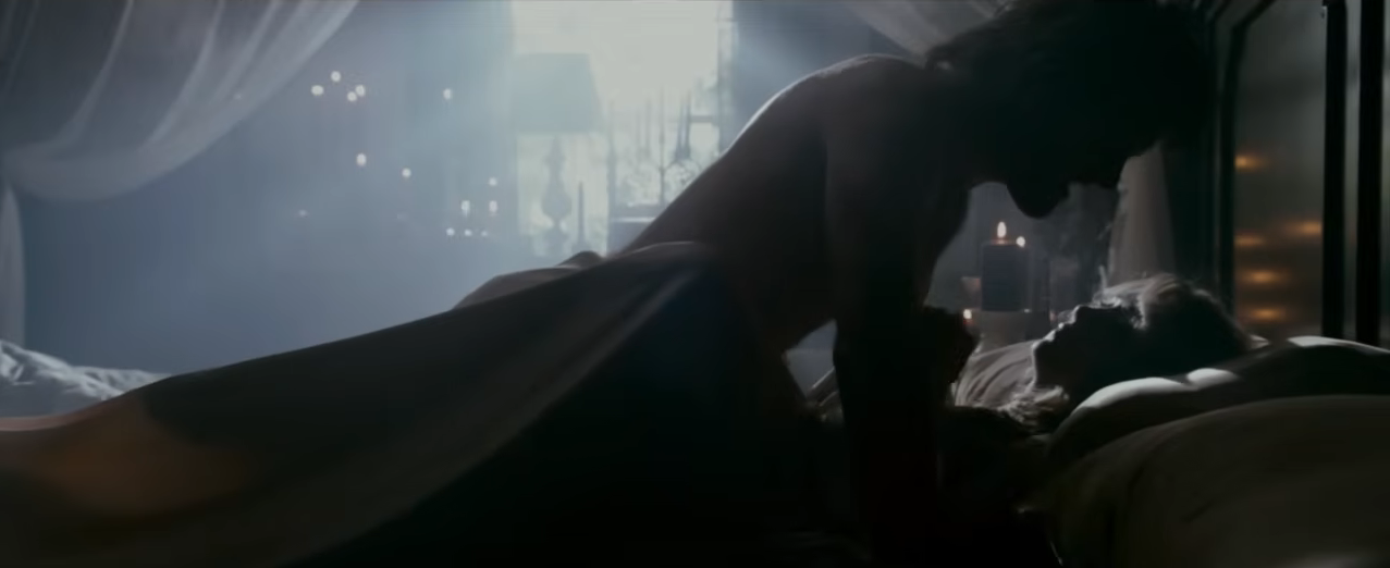 A man and woman lie on a bed in an intimate scene, softly lit. The setting is a dimly lit room with curtains and candles in the background