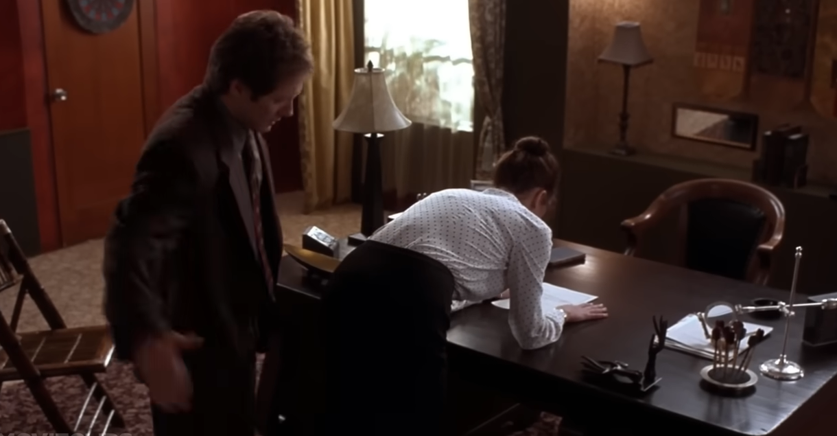 A man in a suit prepares to spank a woman bent over a desk in an office setting