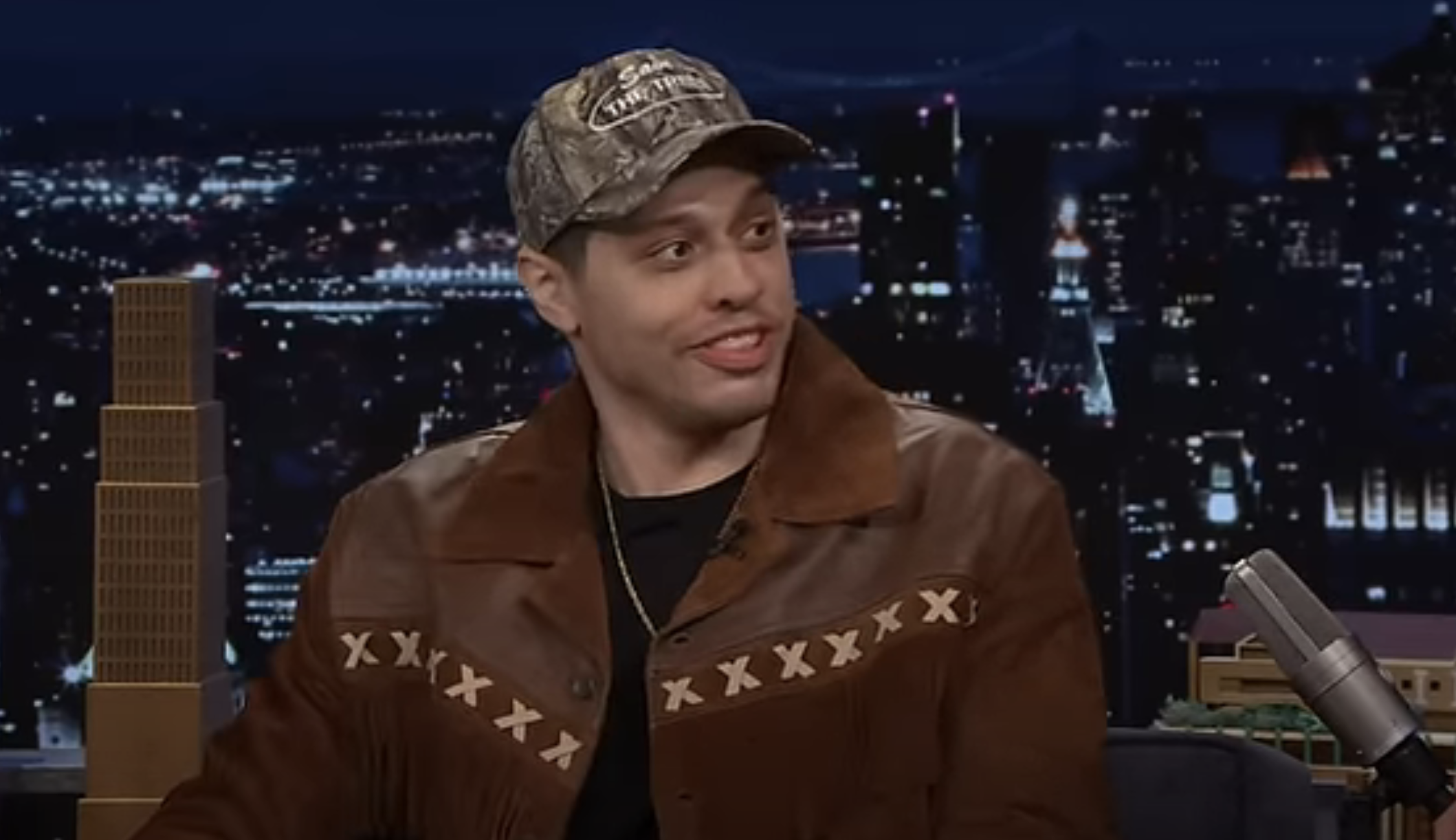 A person in a brown jacket and cap sits in a TV studio with a nighttime cityscape background, engaged in conversation on a talk show