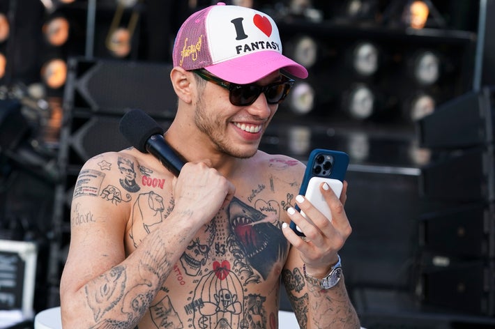 Person with tattoos, wearing sunglasses and a “I ♥️ Fantasy” cap, smiles at phone while holding a microphone
