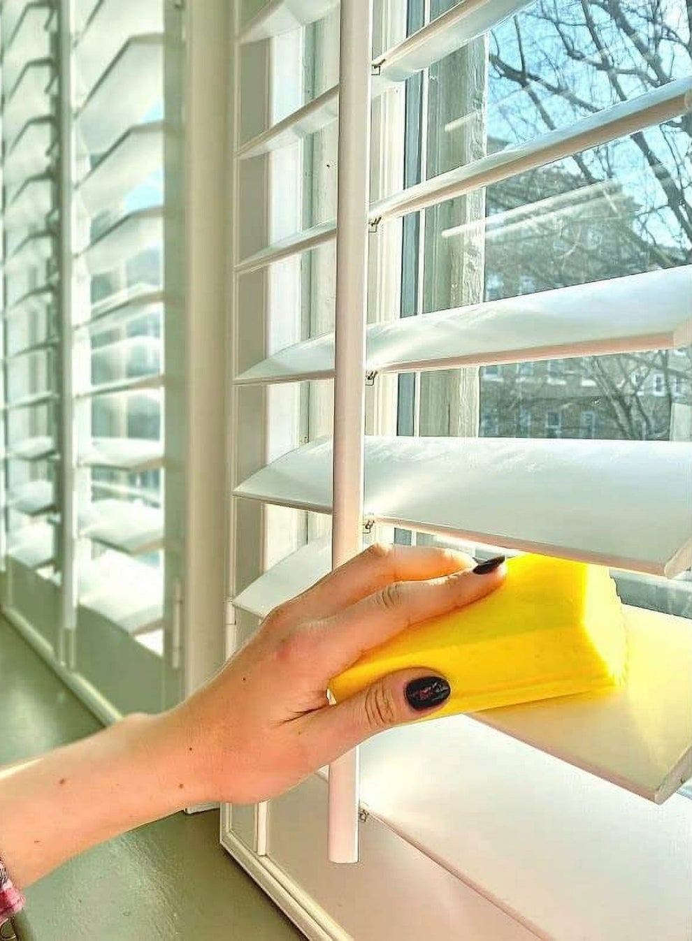 A person cleans white window blinds with a yellow sponge, showcasing cleaning products for home maintenance