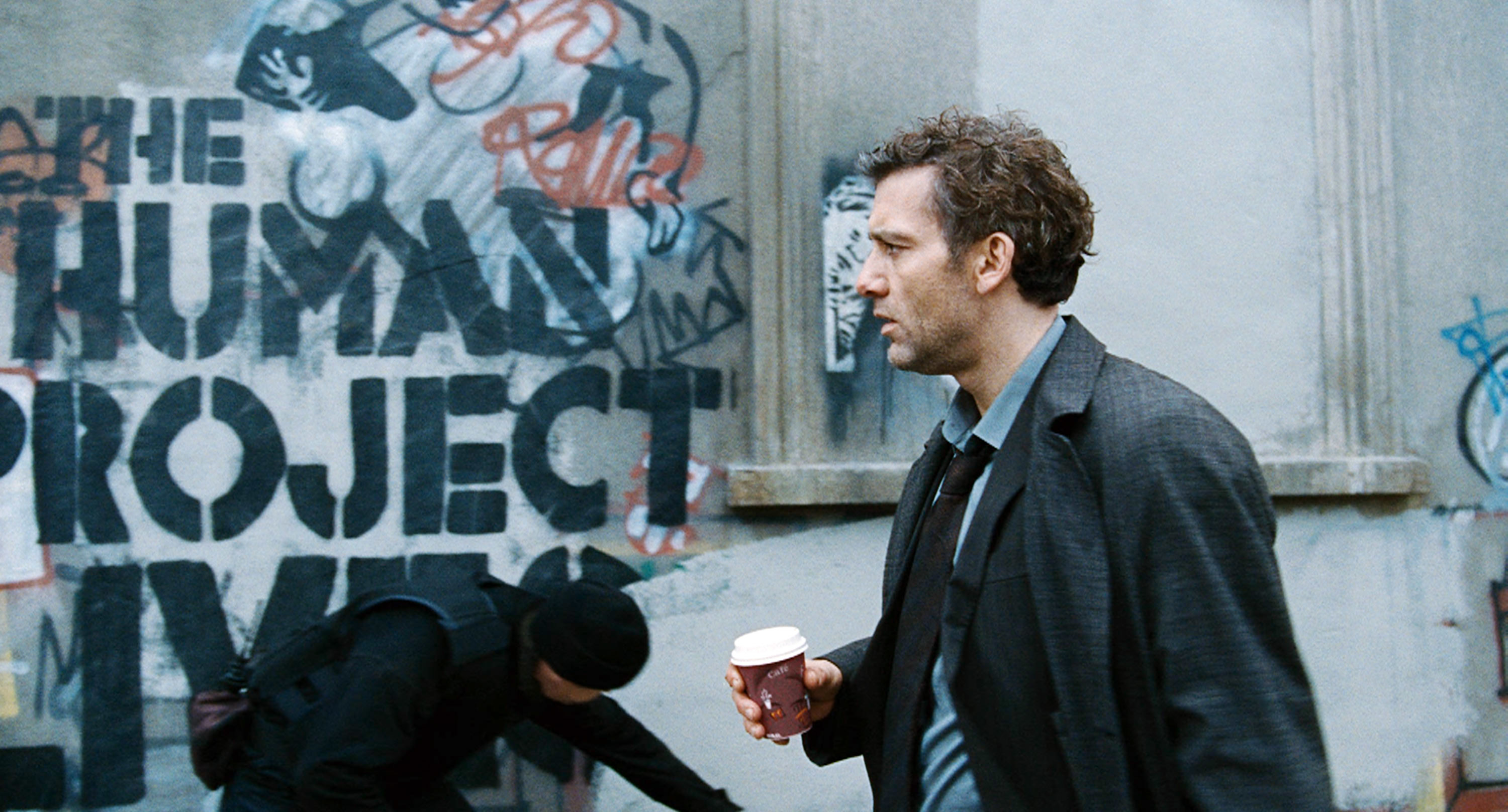 Person holding a coffee cup walking past a graffiti-covered wall that reads "The Human Project Lives." Another person is crouched down
