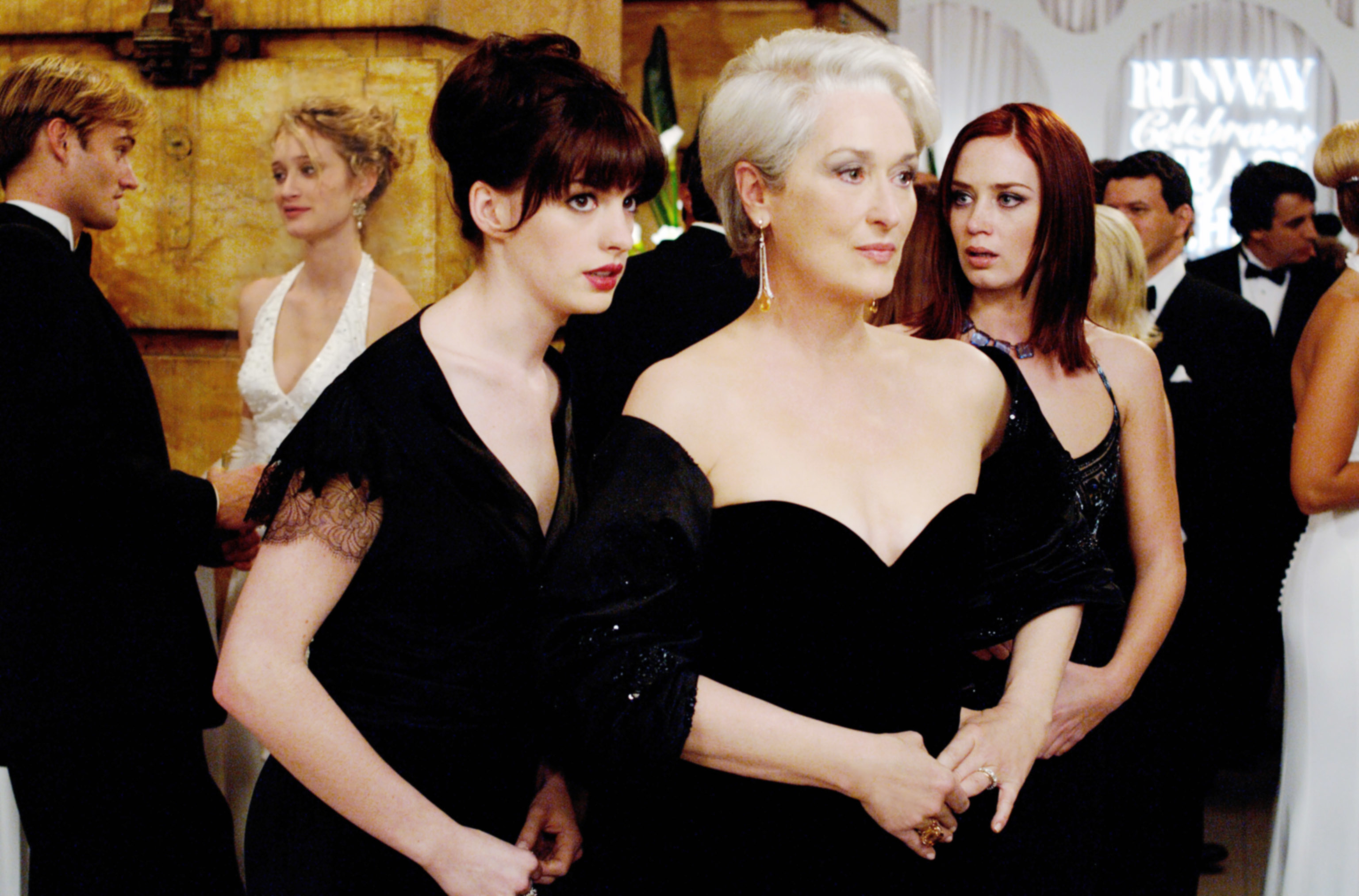 Three women wearing elegant evening gowns at a formal event. They appear to be watching something intently