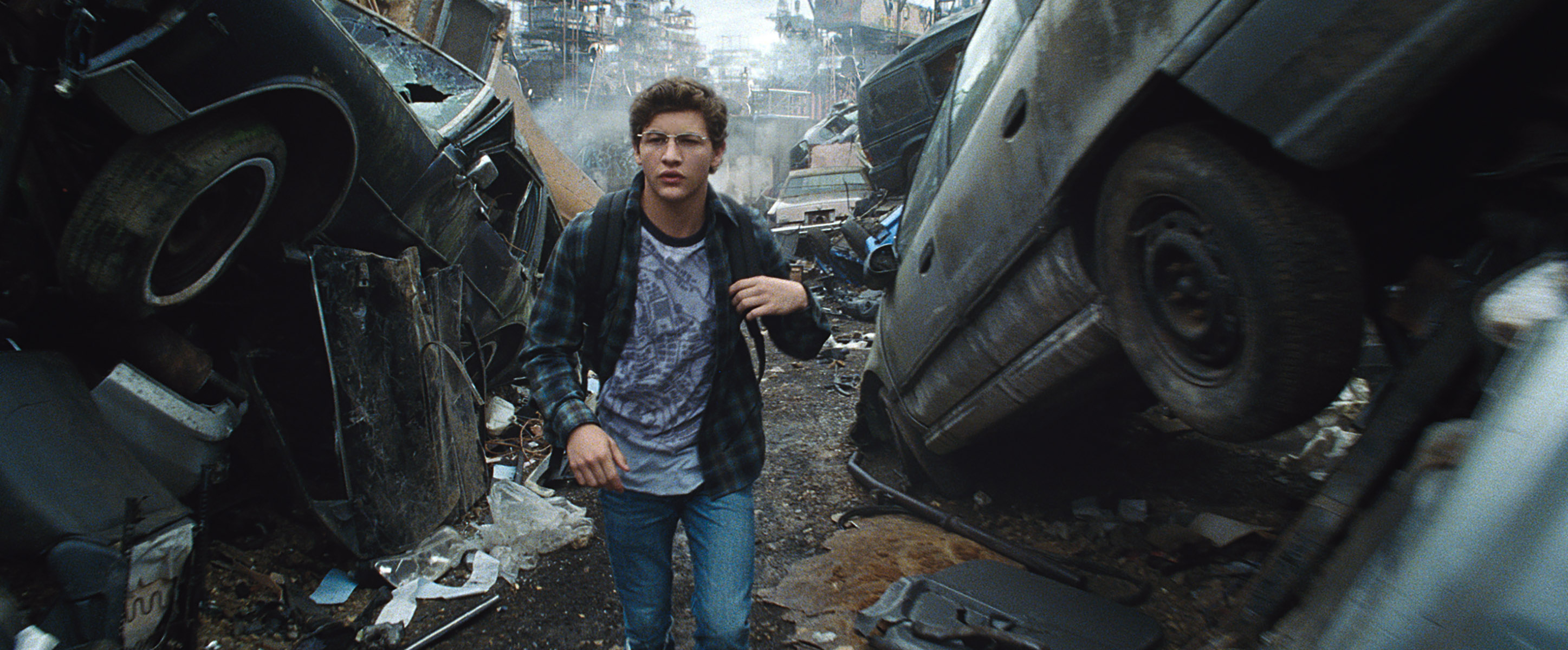 A young man walks through a dystopian scrapyard, wearing a casual t-shirt and jacket, navigating between piles of wrecked vehicles
