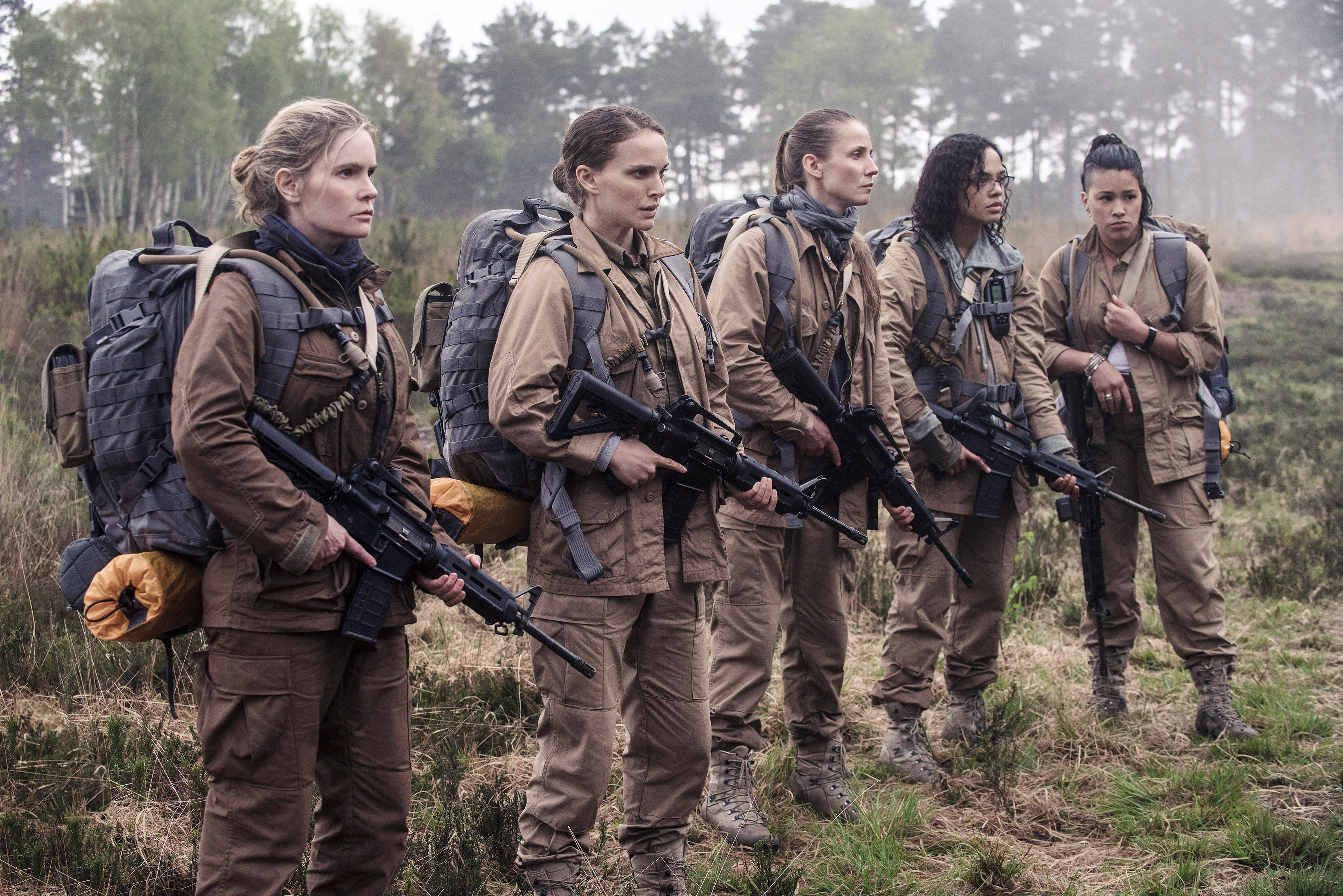 Five people in tactical gear with rifles stand in a field, appearing focused and ready for action