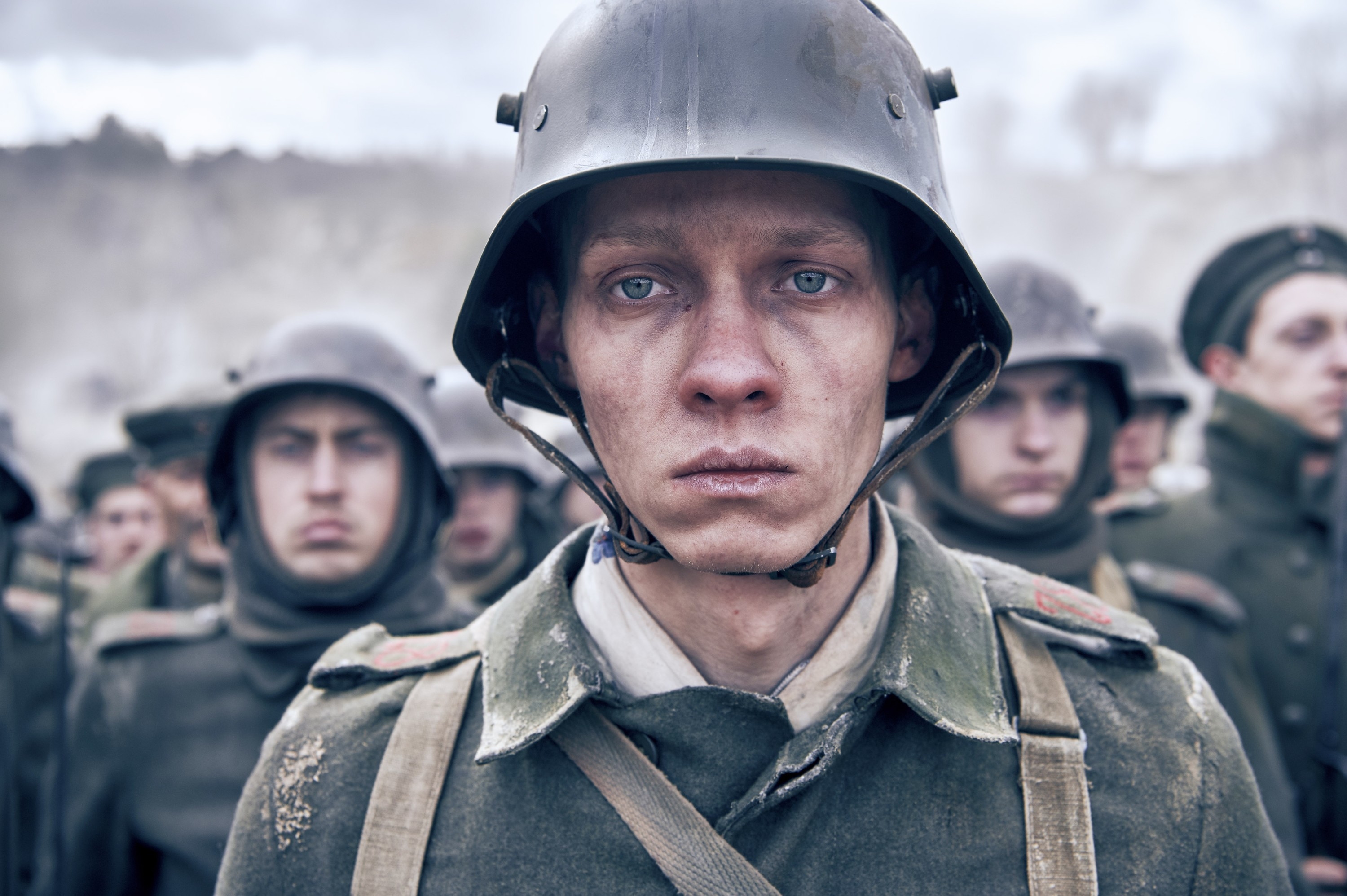 A soldier in a battle-worn uniform and helmet looks directly at the camera, with fellow soldiers blurred in the background