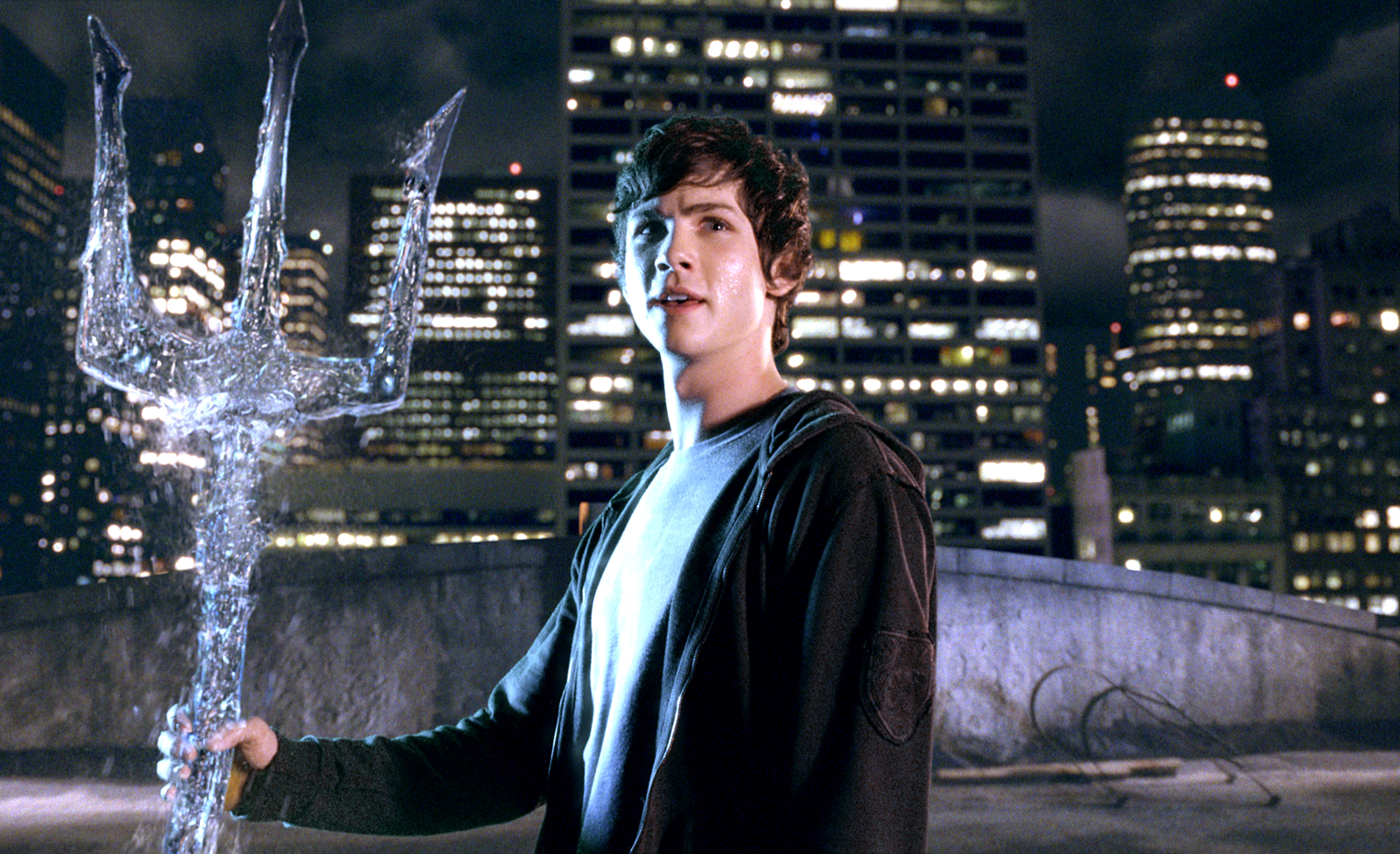 A person stands on a rooftop at night, holding a large trident, with a city skyline in the background