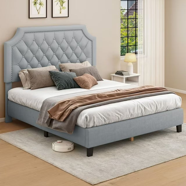 Kamar tidur yang elegan dengan tempat tidur headboard berumbai, berbagai bantal, dan robot vakum di atas karpet, menampilkan dekorasi rumah yang penuh gaya