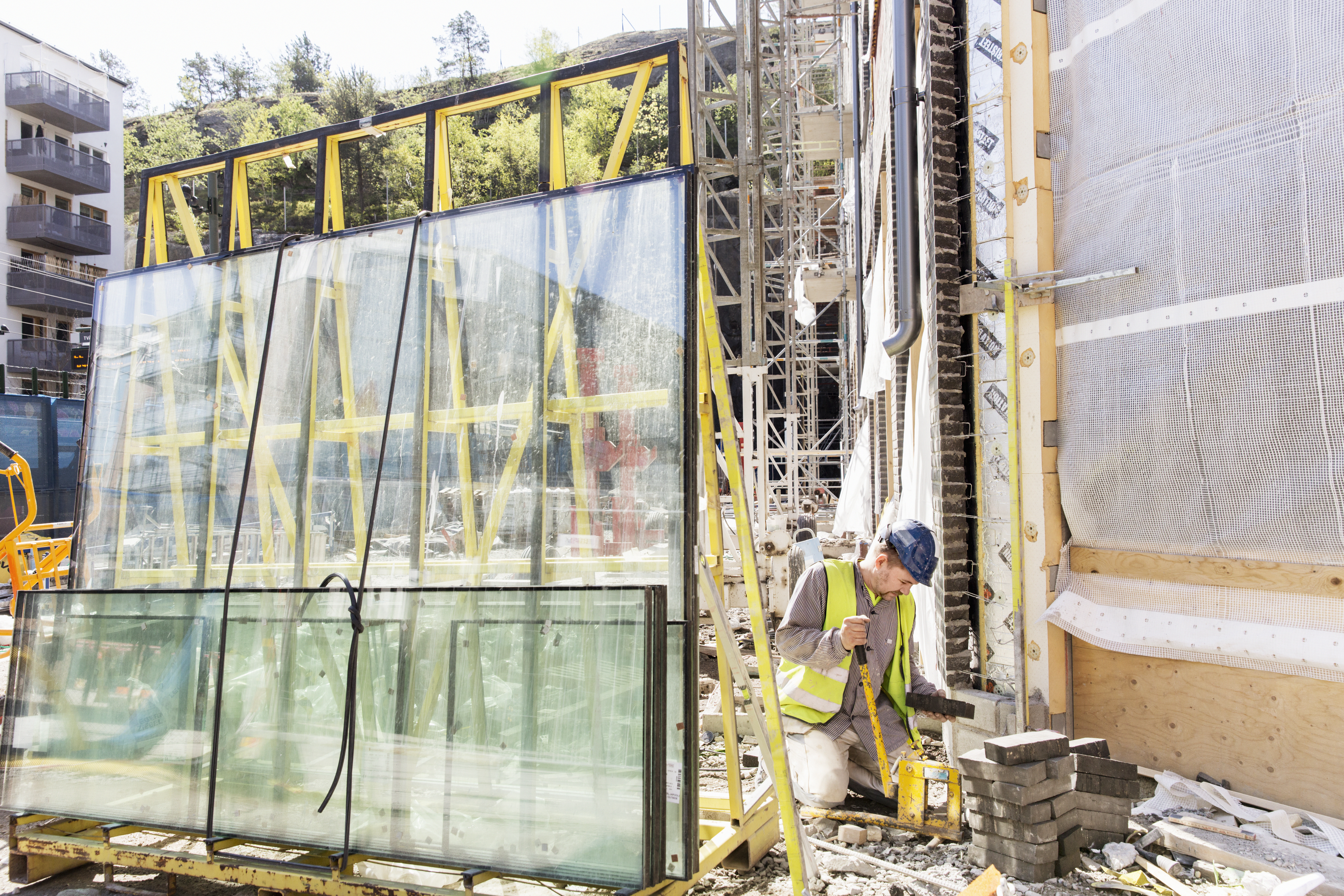 A construction worker in a safety vest and helmet stacks bricks near large glass panels at a building site