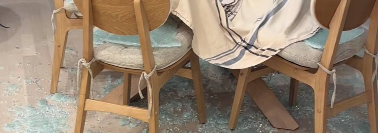 Broken glass table shattered on the floor, surrounded by wooden chairs and a striped cloth