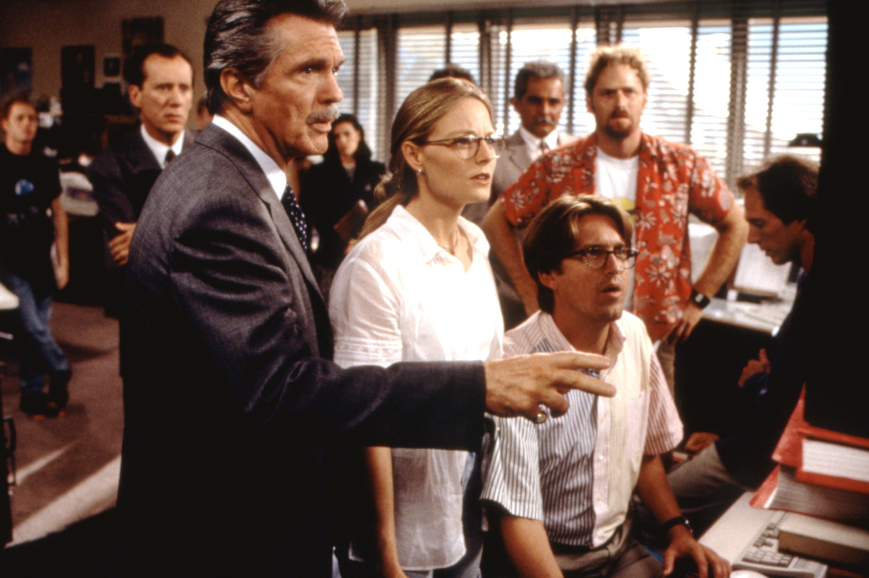A group of people in an office, intensely focused on a computer screen including Jodie Foster; One man points while others stand or sit nearby, watching attentively