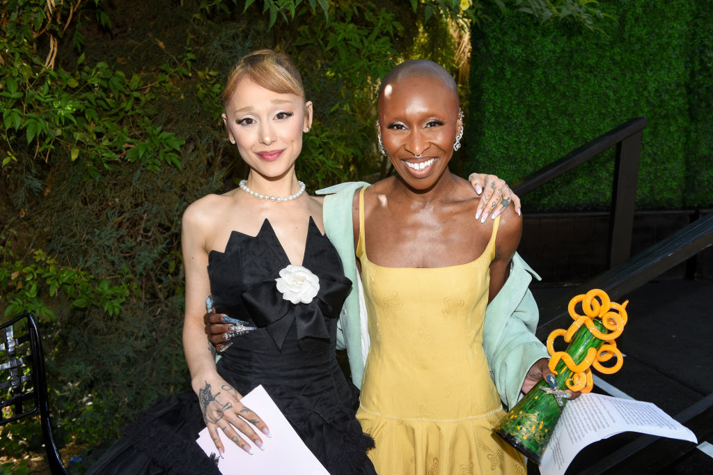 Two individuals pose together, one in a strapless black dress with a white floral detail, and the other in a yellow dress holding a circular-shaped award