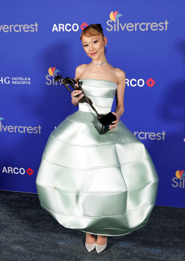 Person in a voluminous, futuristic strapless gown holding an award, standing on a blue carpet at a celebrity event