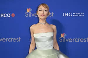 Person in elegant strapless dress on a red carpet, posing in front of a backdrop with logos
