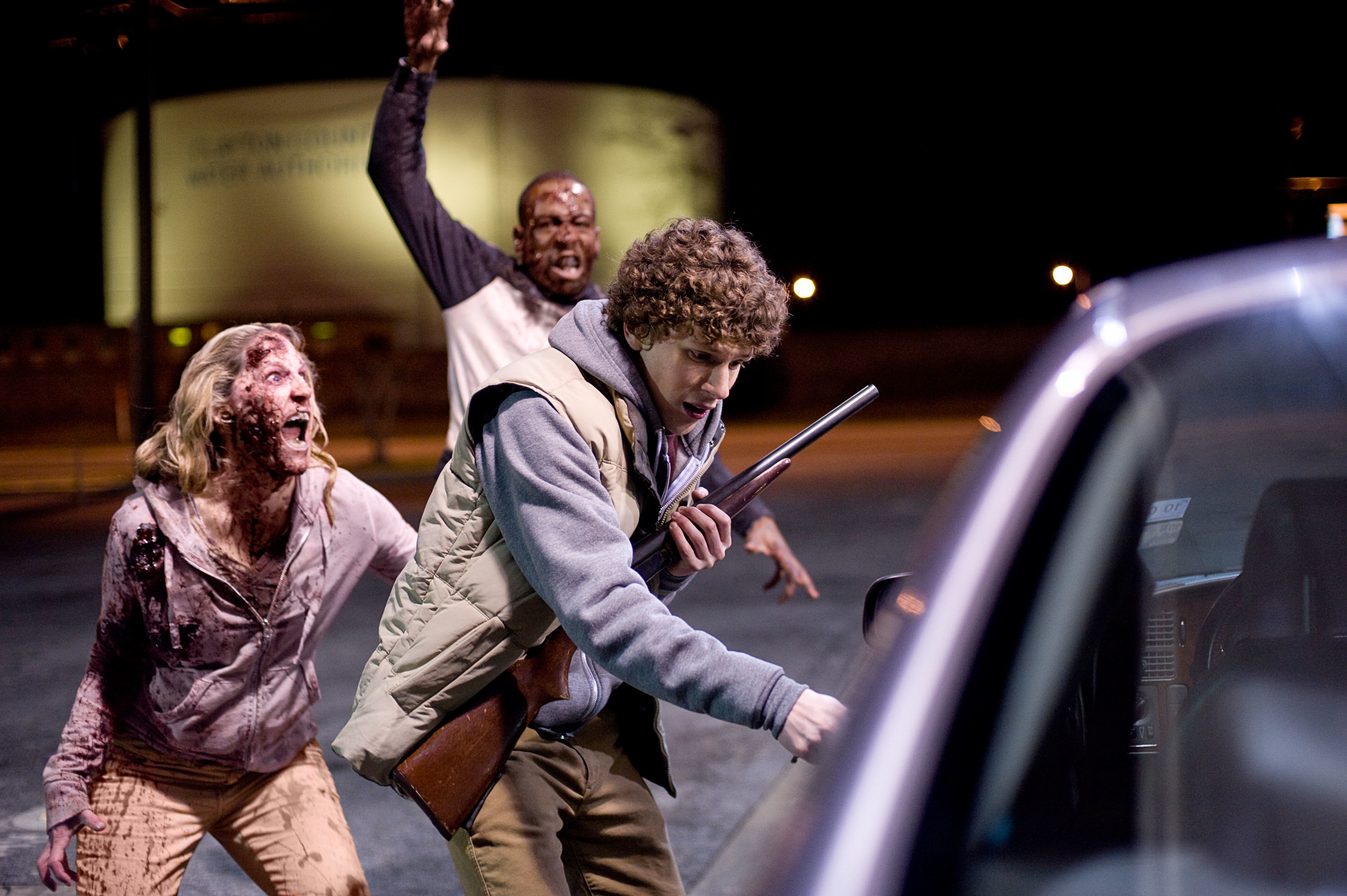 Jesse Eisenberg looks fear stricken and holds a shotgun approaching a car while two zombies follow closely behind