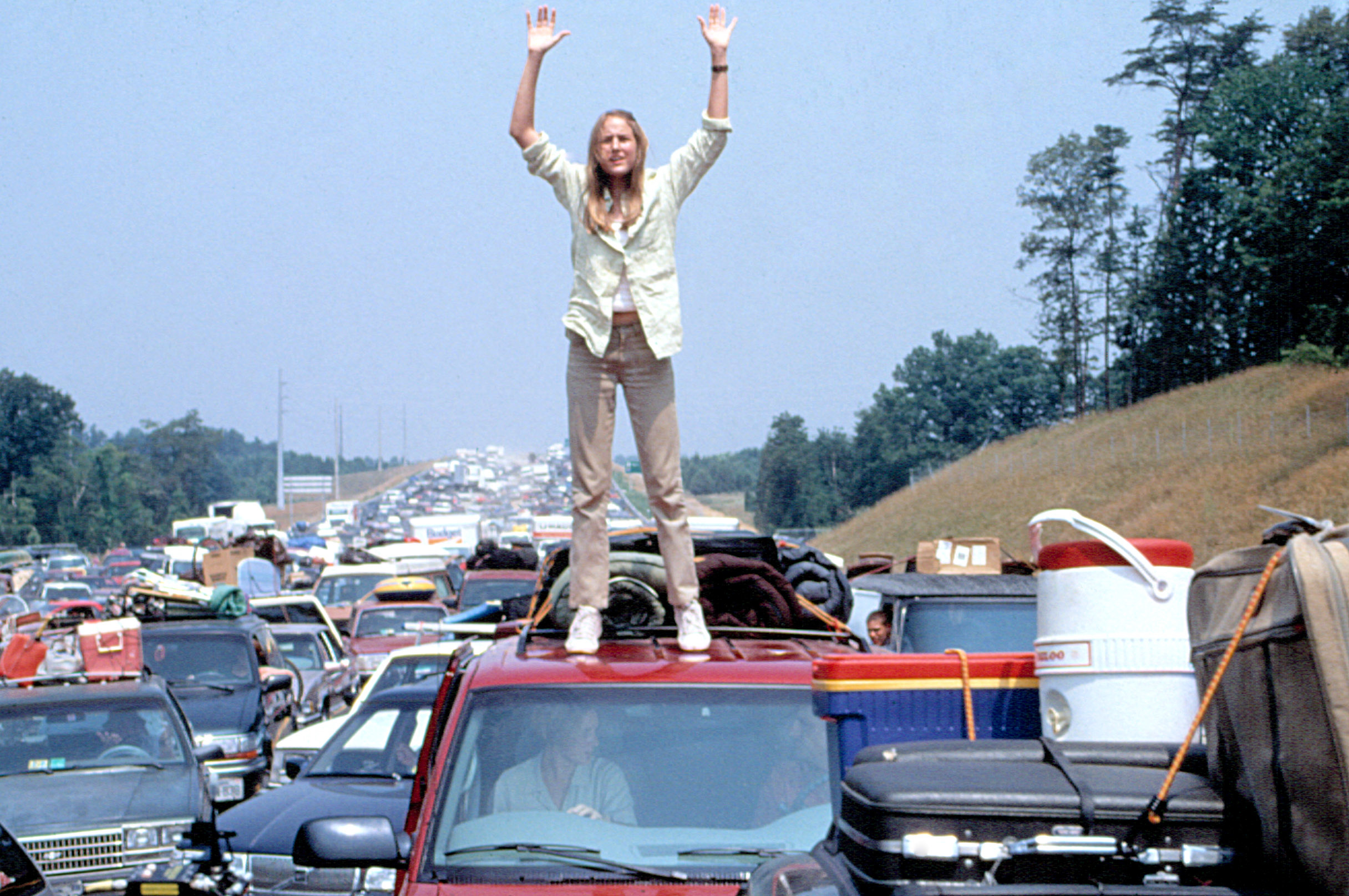 Leelee Sobieski stands on a car roof with hands raised, surrounded by cars in traffic, outdoors