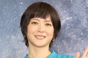 短い髪の人がカジュアルな青いシャツを着て微笑みながら手を振っている。
